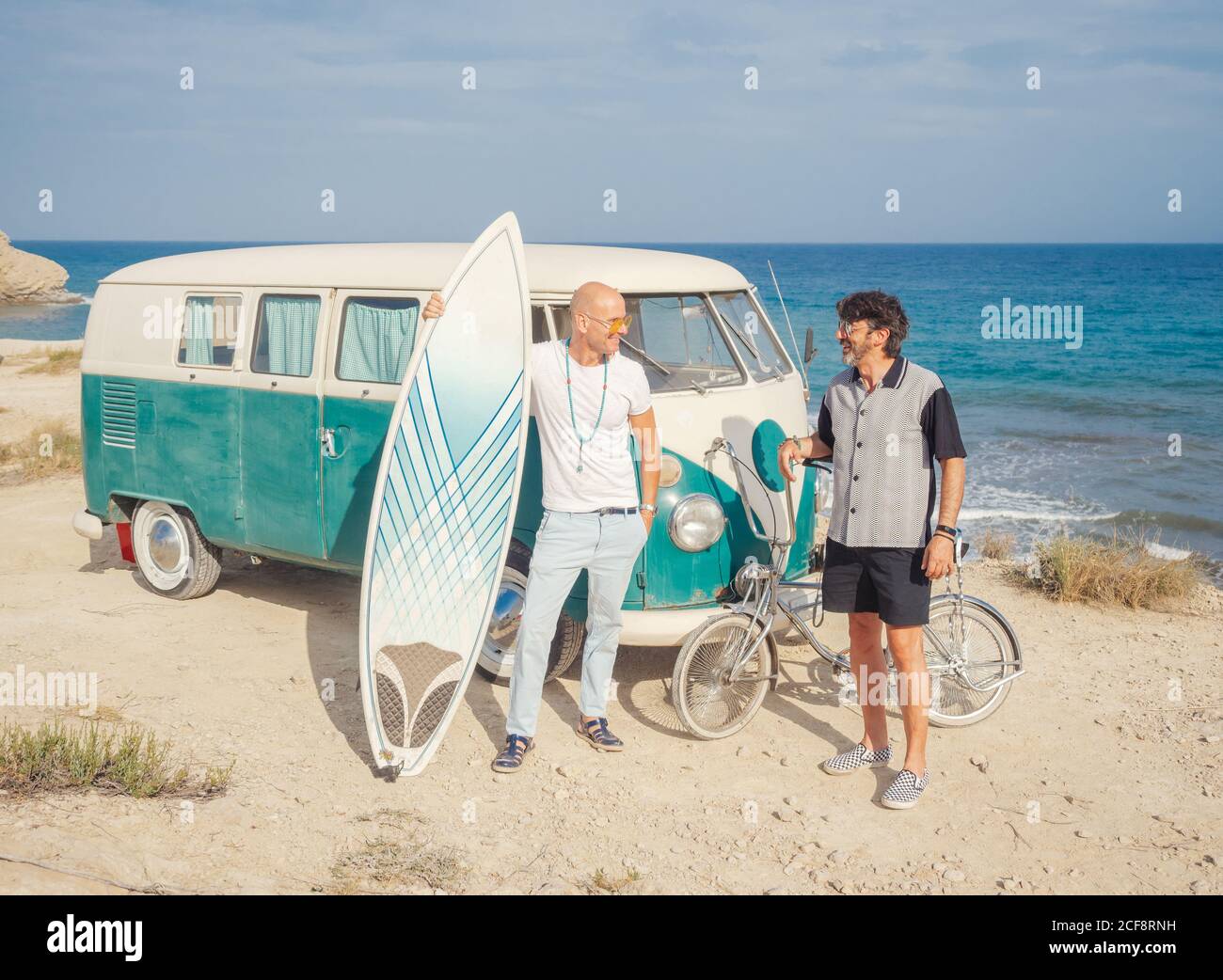 Starke aktive Männer mit großen weißen Surfbrett reden in der Nähe alt Auto und Fahrrad am Meer Stockfoto