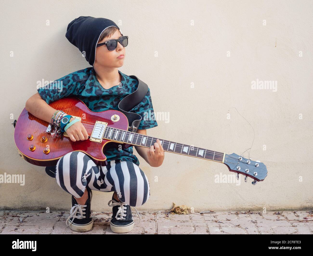 Frech aktiv aufgeregt Junge in bunten Kleidern spielen Gitarre sitzen Lehnend auf Hintergrund der weißen Wand Stockfoto