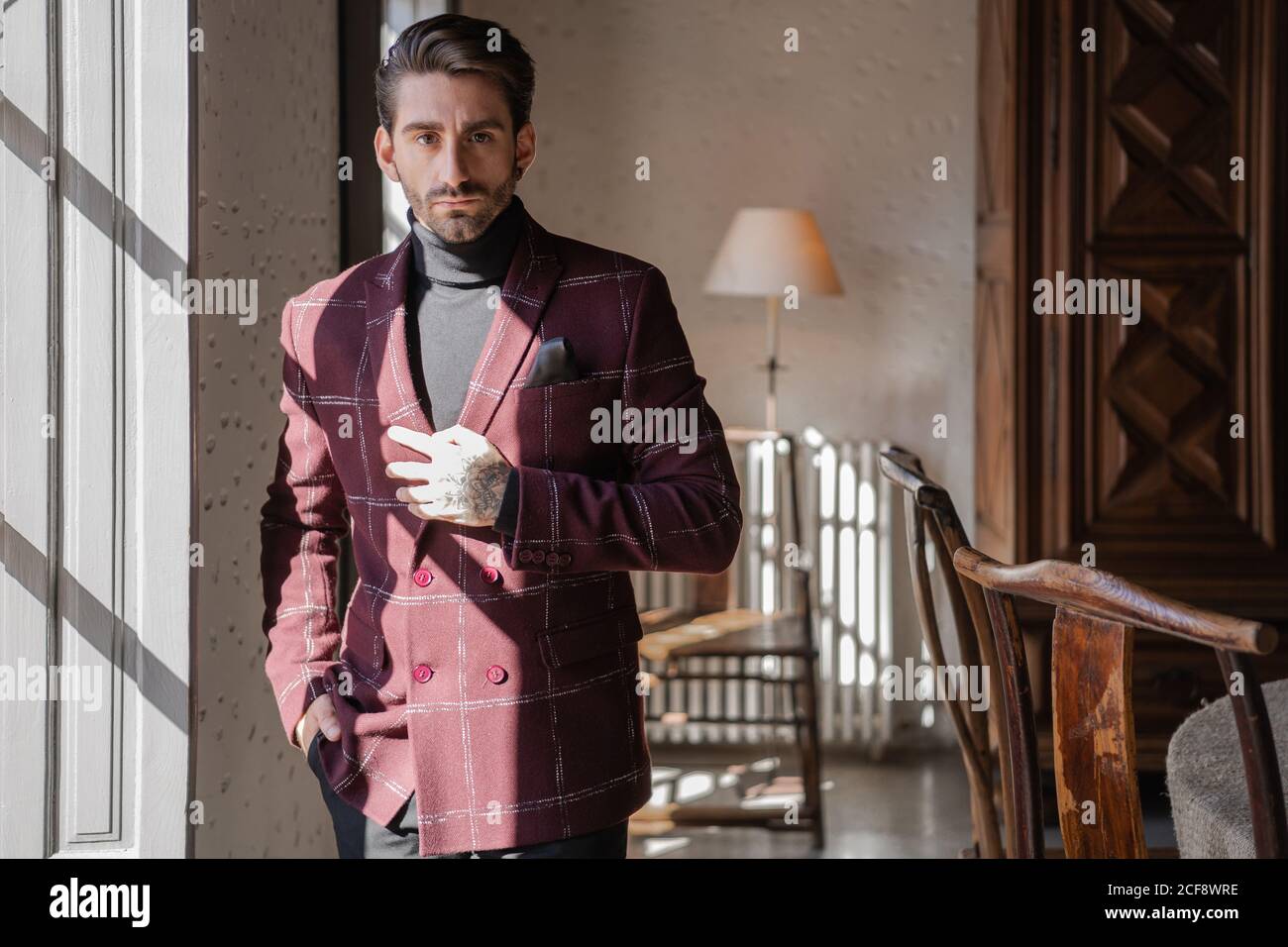 Ruhige pensive Geschäftsmann in karierten Burgunder Jacke mit schwarzem Hemd Und Taschentuch Blick auf die Kamera und treffen harte Entscheidung Beim Stehen gegen verschwommene Vintage-Interieur mit antiken Möbeln und Grün gezeichnete Baum im geräumigen Badezimmer Stockfoto