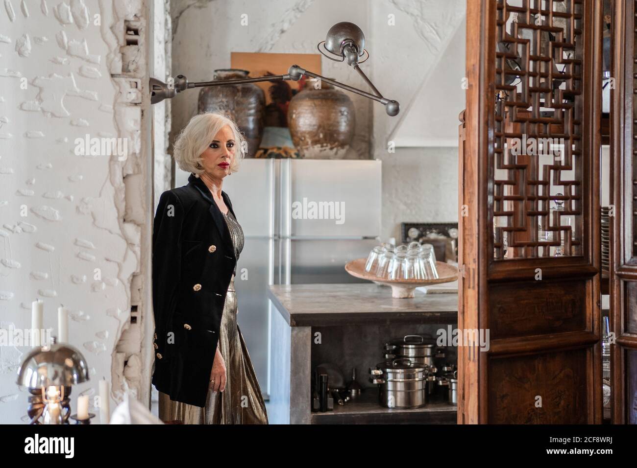 Ernst selbstbewusst weiblich in eleganten schwarzen Jacke und goldenes Hemd Und die Hose schaut weg und kontempliert, während sie mit der Hand steht An der Hüfte neben Holztür gegen verschwommenes Kücheninterieur an Zu Hause Stockfoto
