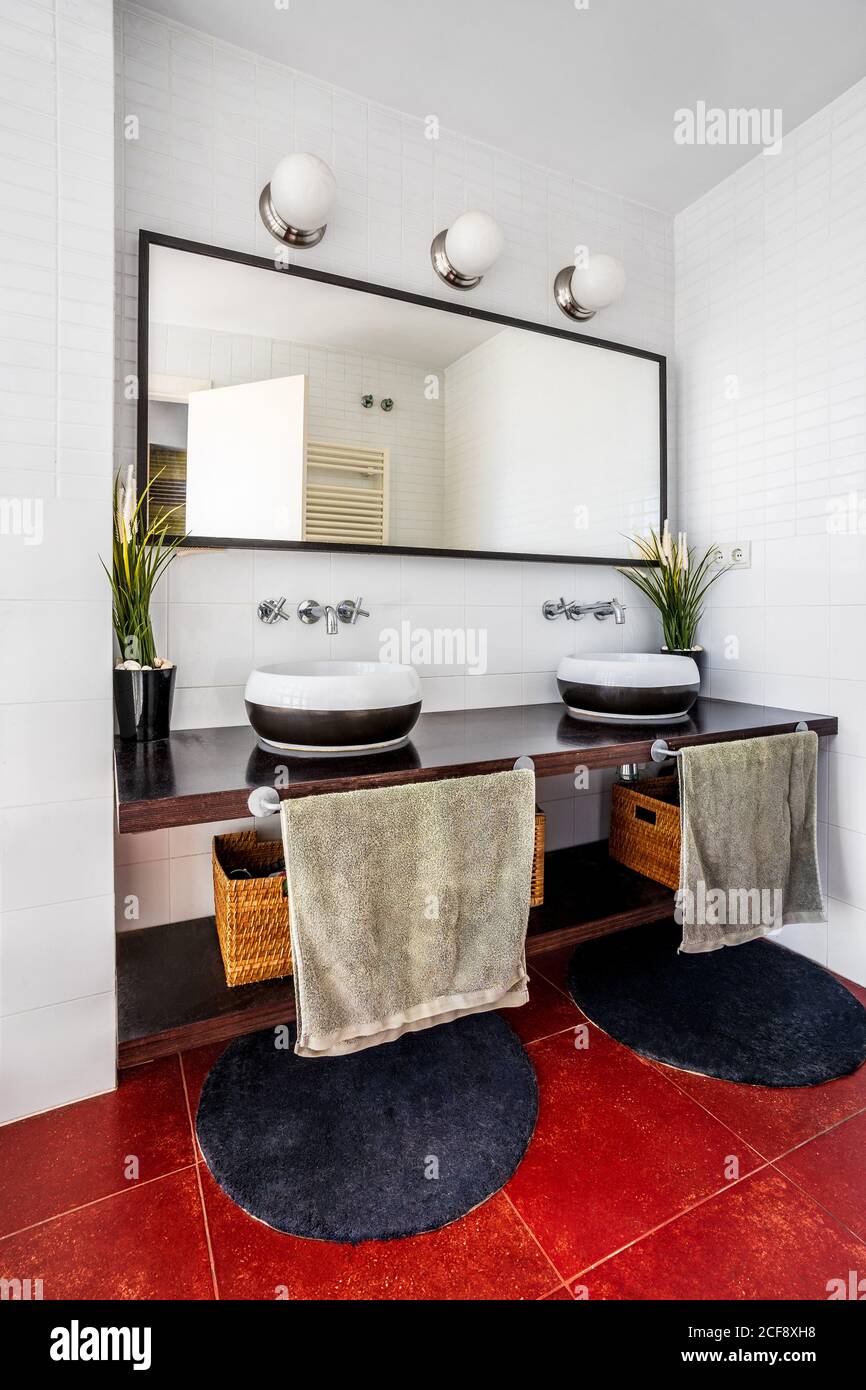 Modernes Bad mit Lampen und großem Spiegel über stilvoll platziert Theke mit Waschbecken und gemütlichen runden Teppichen auf rotem Boden Stockfoto