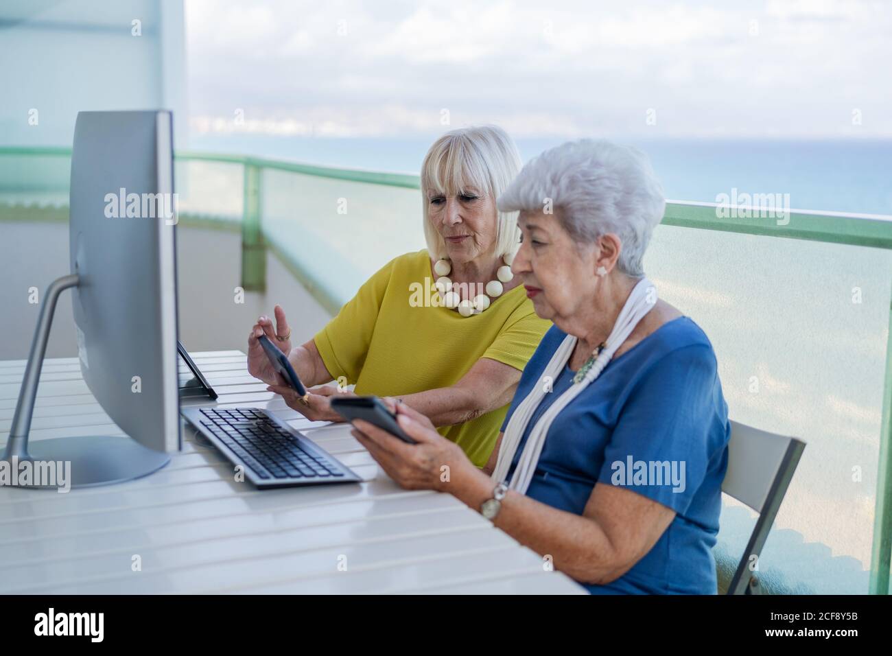 Stilvolle alte Freunde, die während des Sitzens in sozialen Medien auf dem Tablet surfen Am Tisch mit Computer auf dem Balkon im Resort Stockfoto