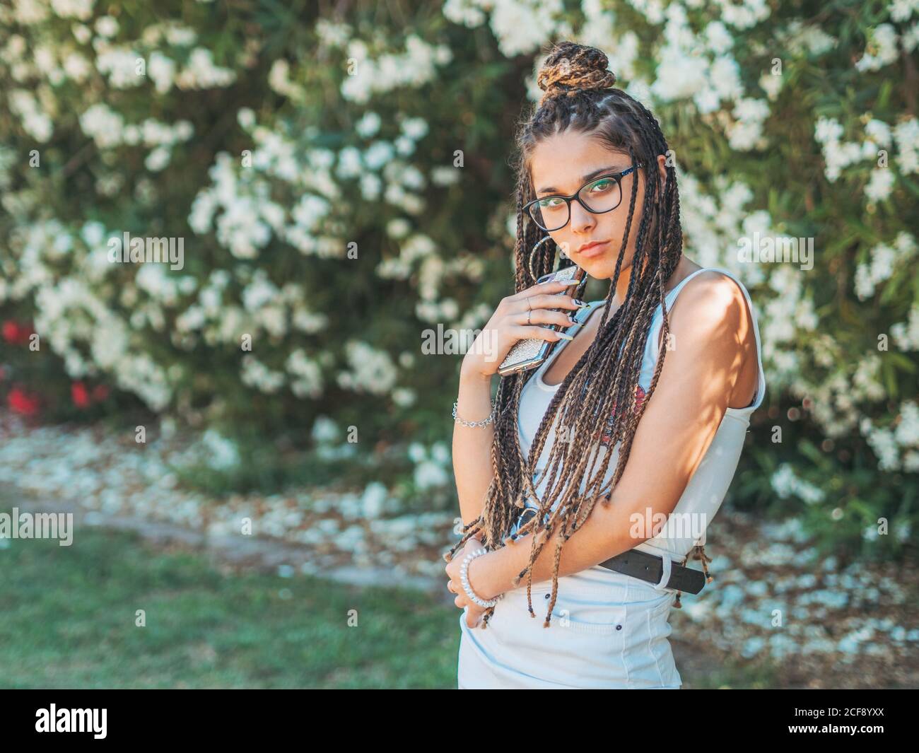 Stilvolle junge Frau trägt Brille Dreadlocks und Sommerkleidung mit Smartphone und Blick auf die Kamera Stockfoto