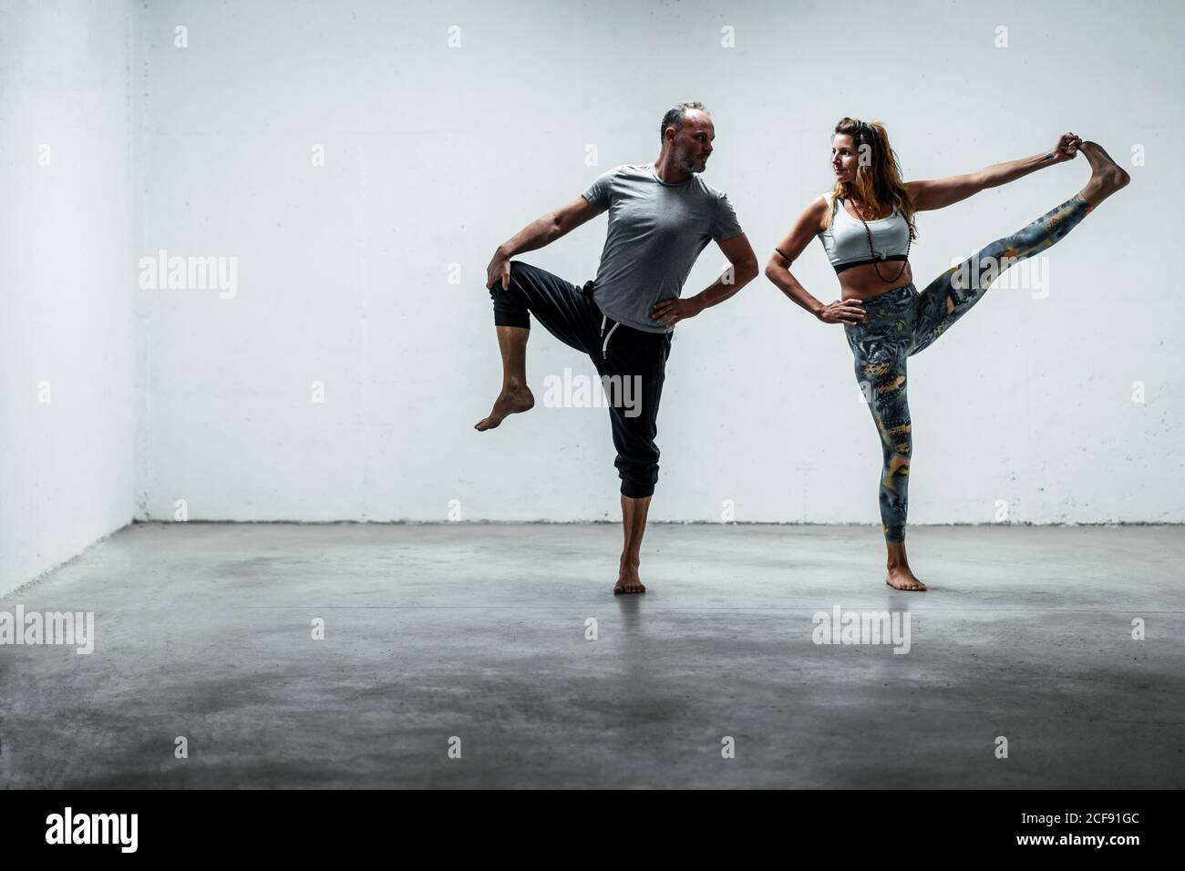 Erwachsenes Paar in aktiver Kleidung, das barfuß auf Betonboden steht In der Hand zu Toe Asana und einander anschauend Beim Balancieren auf dem Bein Stockfoto