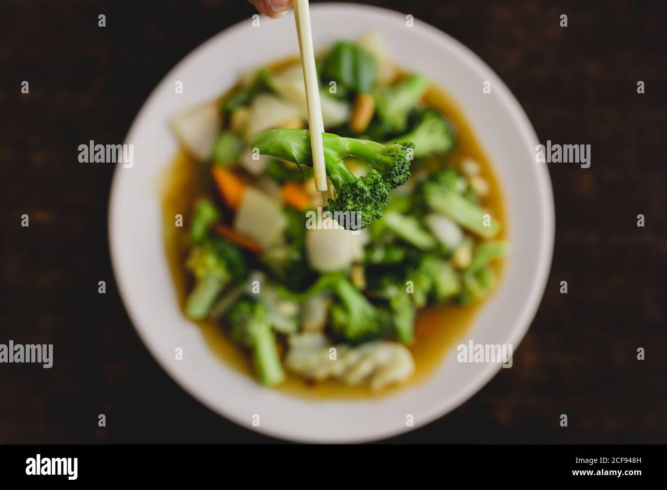 Von oben Hand hält leckere Brokkoli mit Essstäbchen über Suppe mit Karotte, Zwiebel und Paprika in asiatischen Restaurant Stockfoto