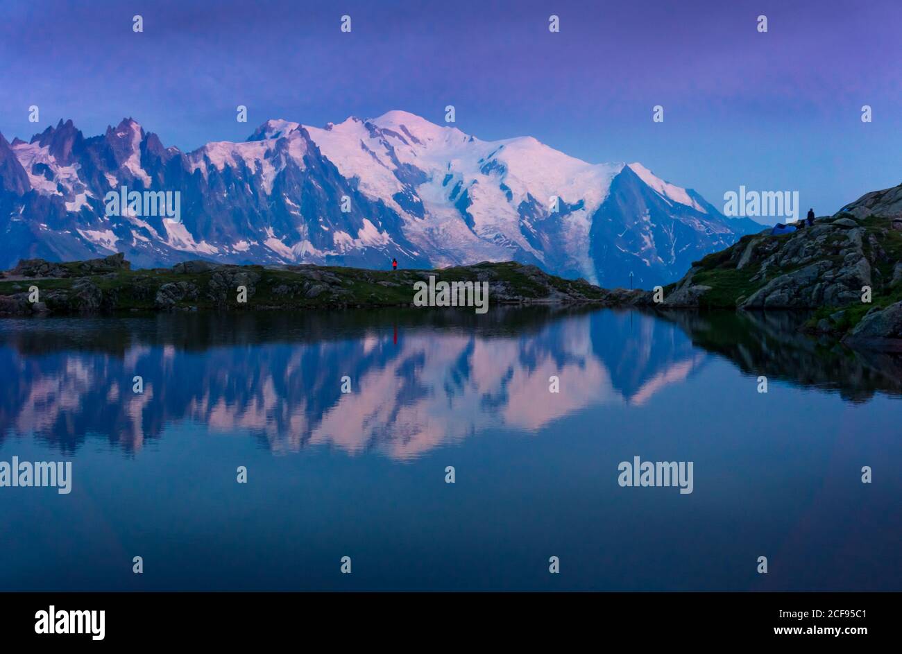 Einsamer Tourist auf hügeligen Ufer spiegelt sich in kristallklarem See in Schneebedeckte Berge im Sonnenlicht Stockfoto