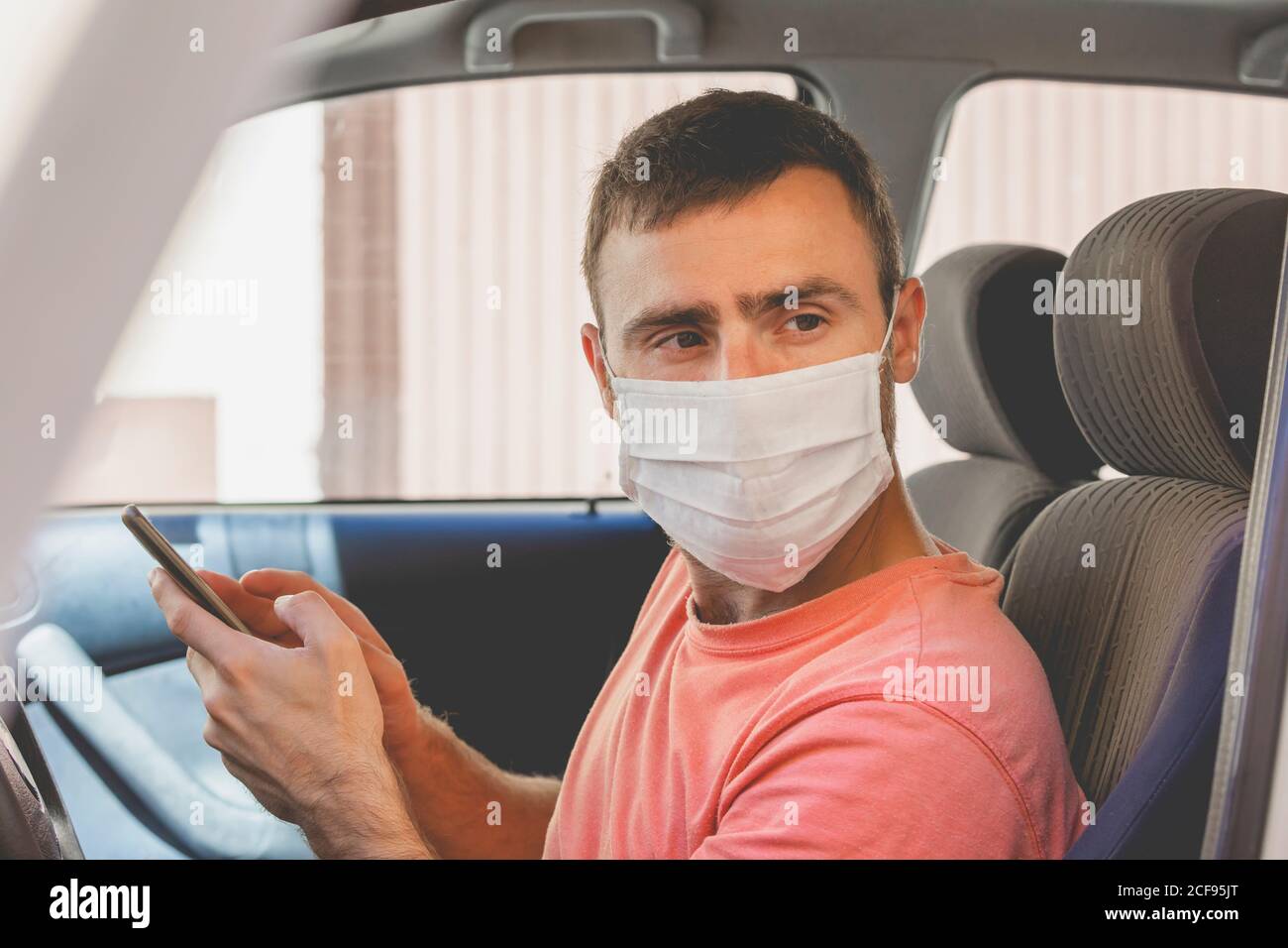 Ein junger Mann, der eine Stoffmaske trägt, überprüft das Handy im Auto, während er vor der Fahrt in Spanien auf jemanden wartet. Stockfoto