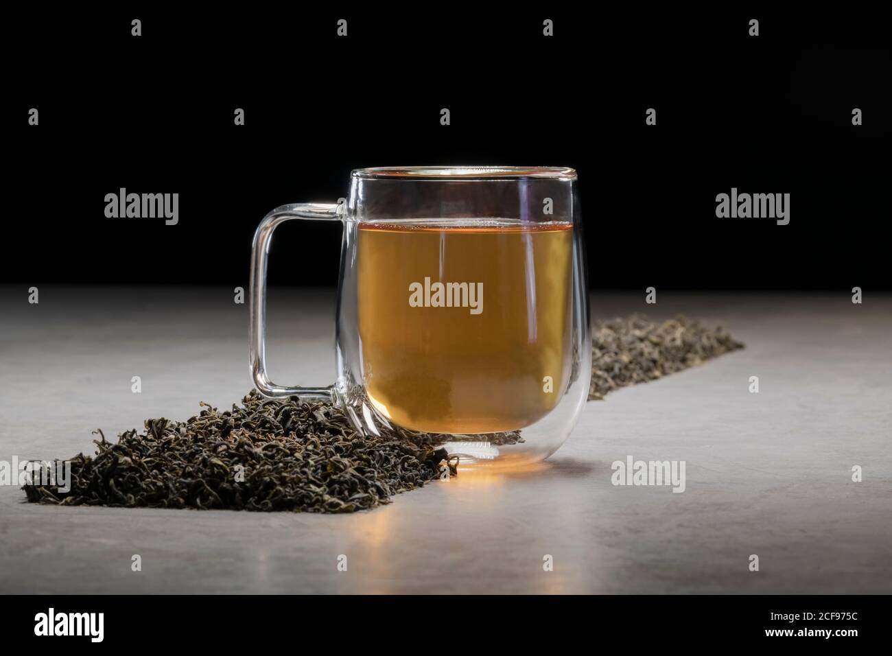 Aromatisches Getränk in Glas Becher mit Haufen von getrockneten angeordnet Teeblätter auf dem Tisch auf schwarzem Hintergrund Stockfoto