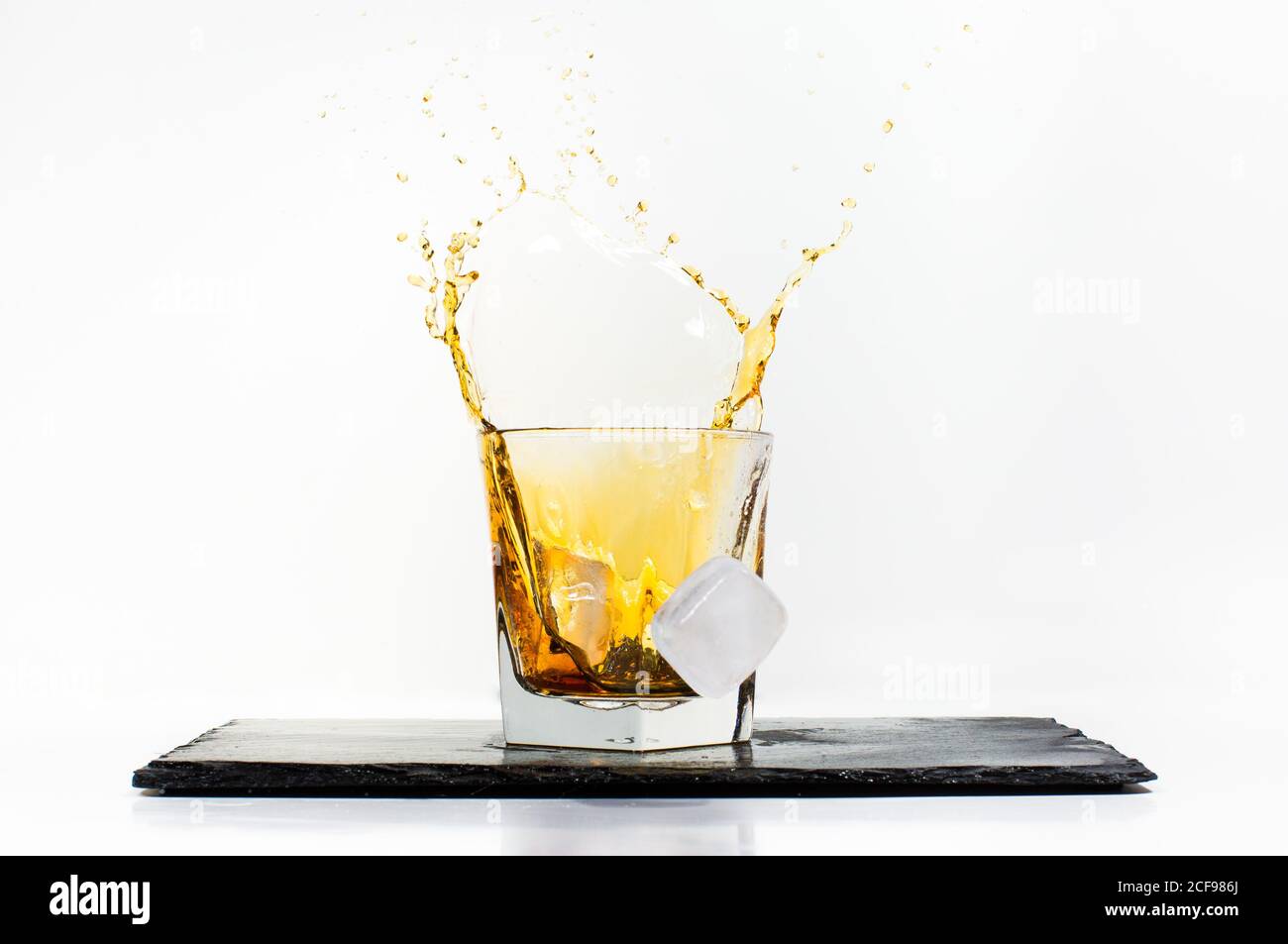 Glas gelbes kaltes Getränk mit fallendem Eiswürfel und Spritzer auf schwarzem Schiefertafel auf weißem Hintergrund platziert Stockfoto