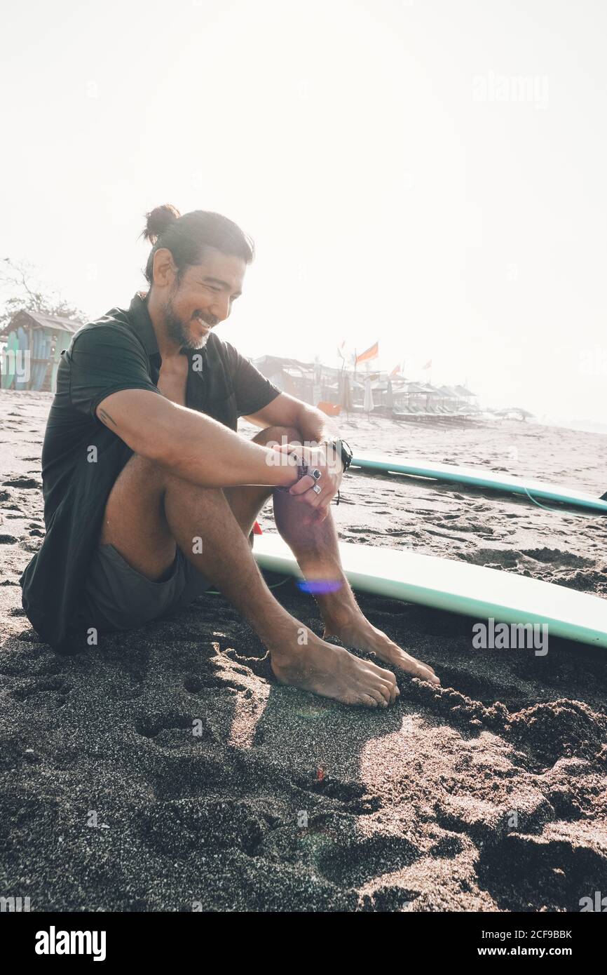 Positive junge ethnische männlich in unknöpfbaren Hemd und Shorts genießen Sommertag, während Sie sich am Sandstrand in der Nähe von Surfbrett ausruhen Stockfoto