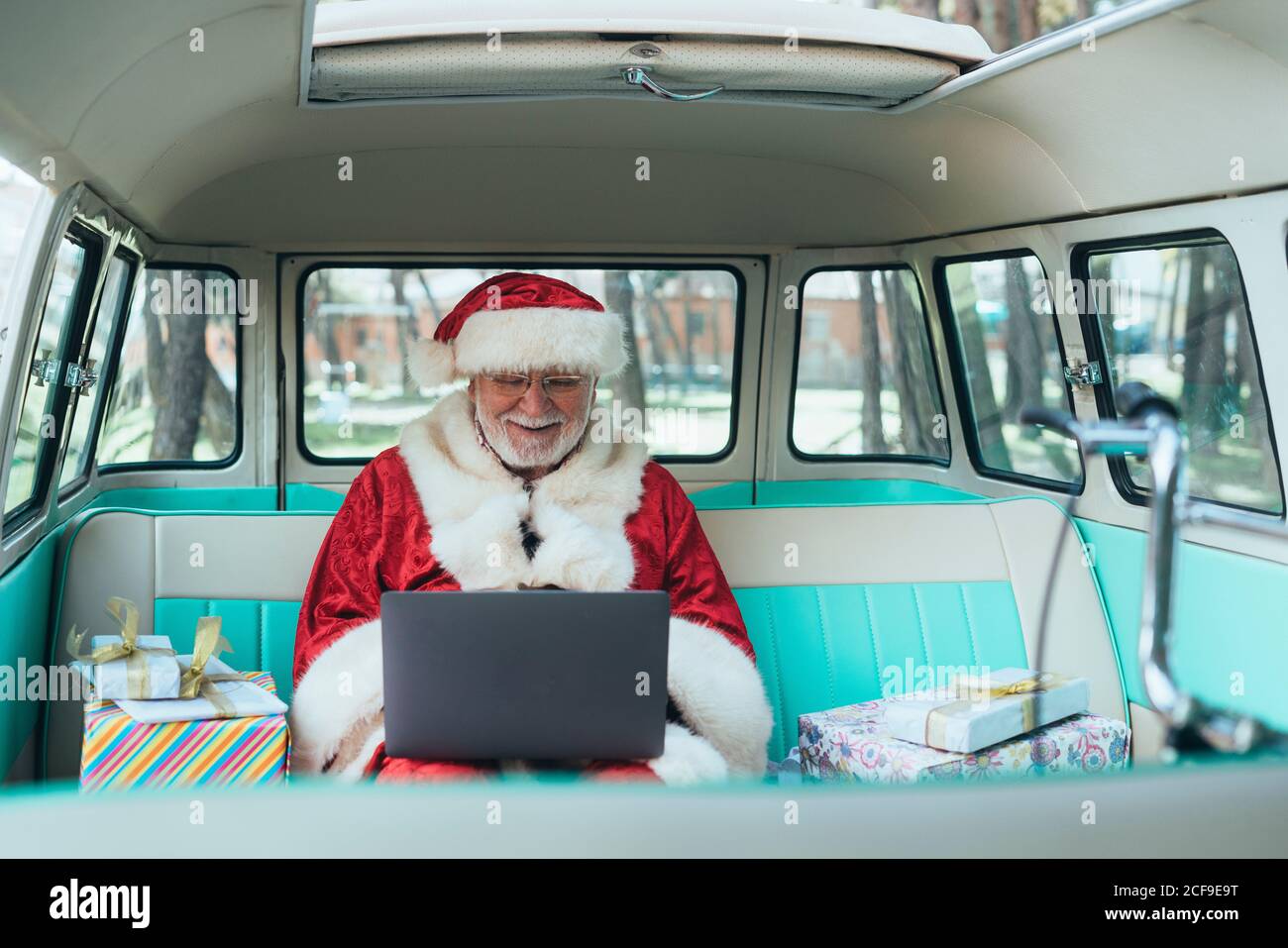 Fröhlicher Mann im Kostüm des Weihnachtsmannes, der im Van sitzt Mit Laptop und bunten Geschenken an sonnigen Tagen Stockfoto