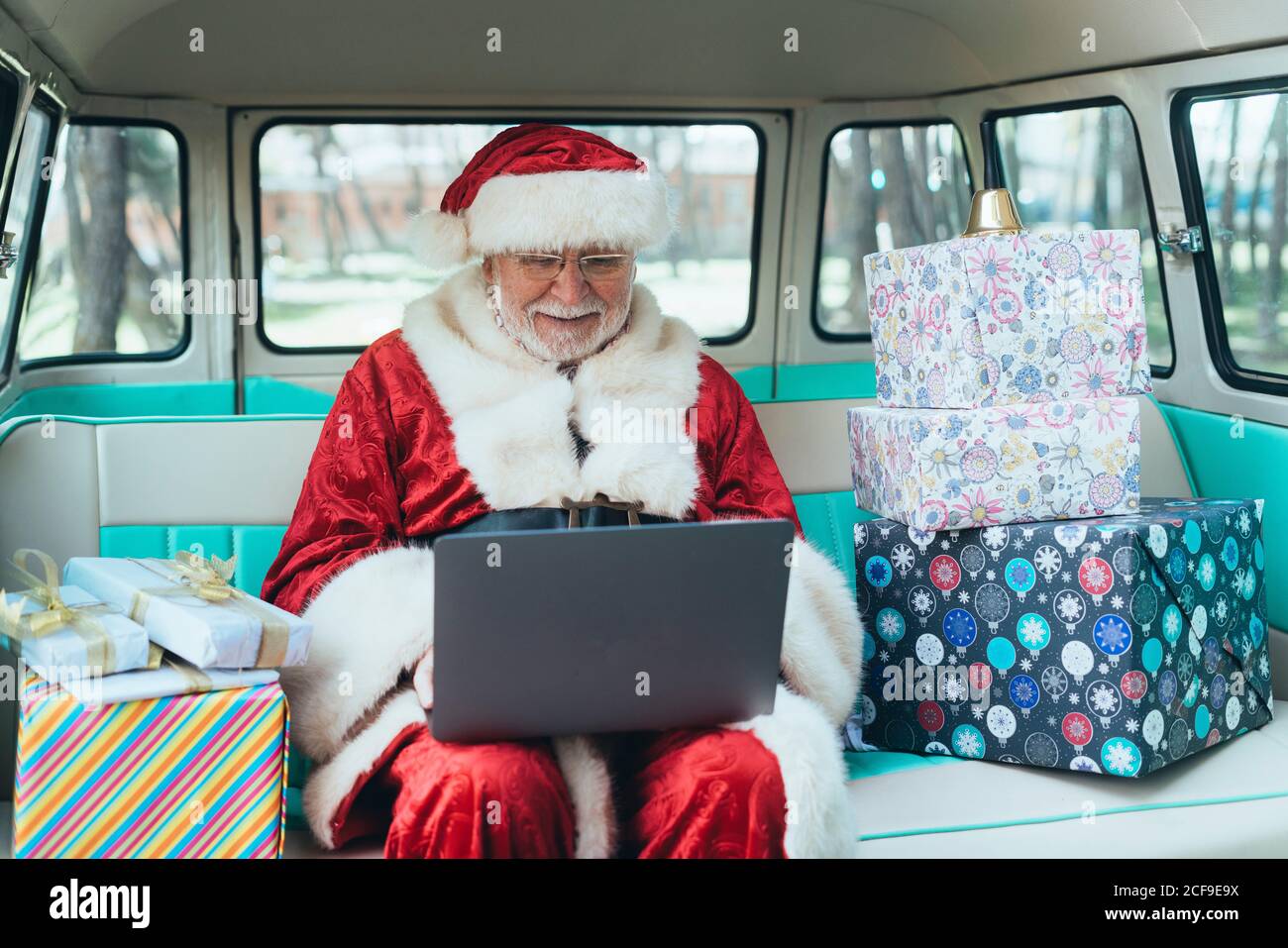 Fröhlicher Mann im Kostüm des Weihnachtsmannes, der im Van sitzt Mit Laptop und bunten Geschenken an sonnigen Tagen Stockfoto