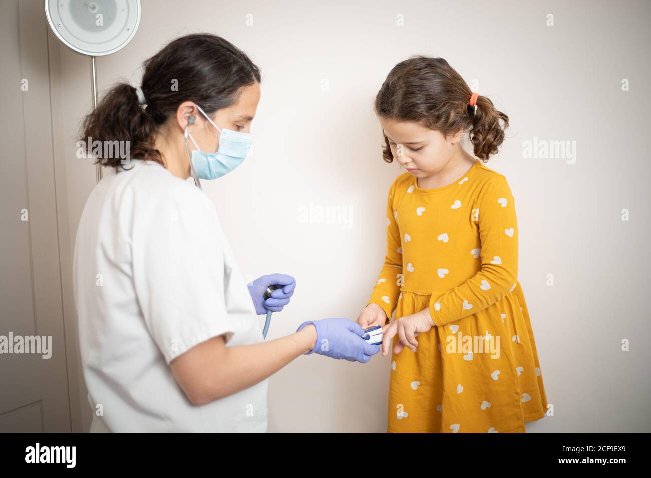 Ein Arzt in Latexhandschuhen mit Fingermanschette zu untersuchen Blutdruck von kleinen Mädchen in lässigen gelben Kleid Stockfoto
