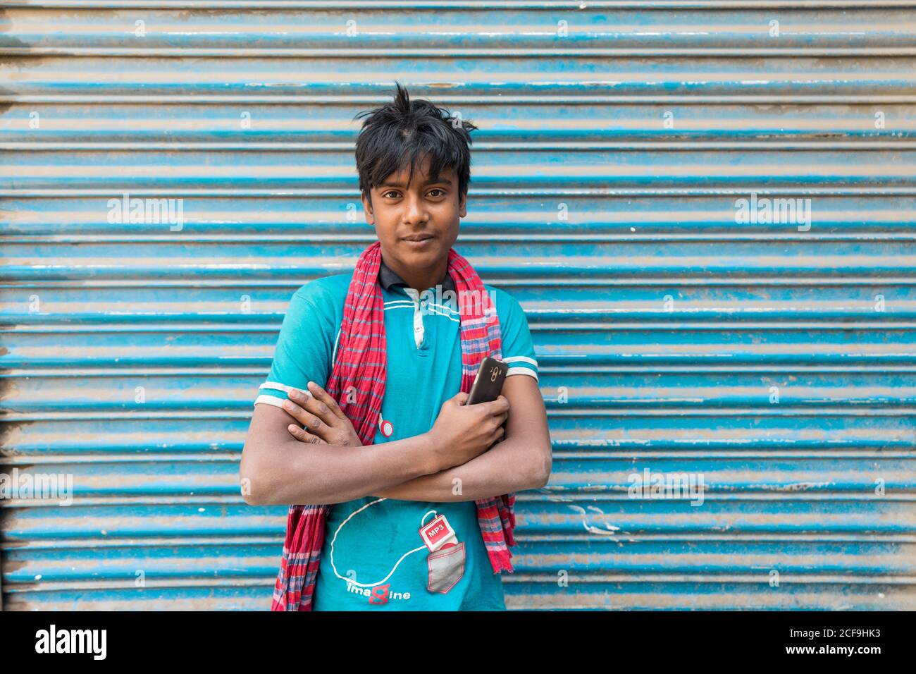 Bangladesch - Januar, 30 2019: Positiver ethnischer Teenager mit Smartphone, das die Arme kreuzt und die Kamera anschaut, während er in der Nähe einer schäbigen Gebäudewand auf der Stadtstraße steht Stockfoto