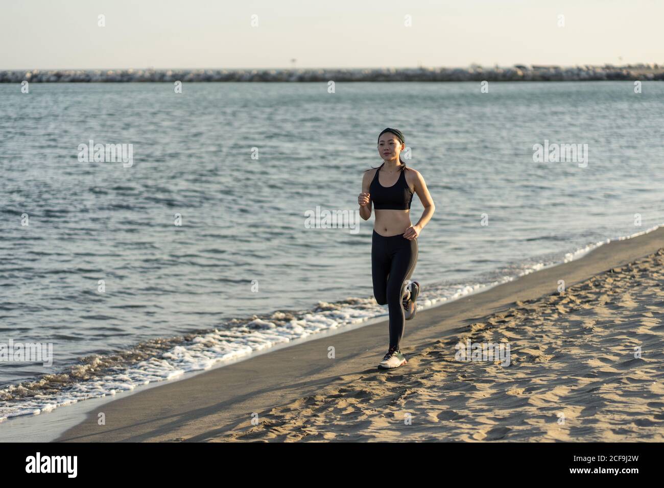 Motivierte junge Sportlerin in aktiver schwarzer Kleidung und Sneakers Joggen entlang der sandigen leeren Küste Stockfoto