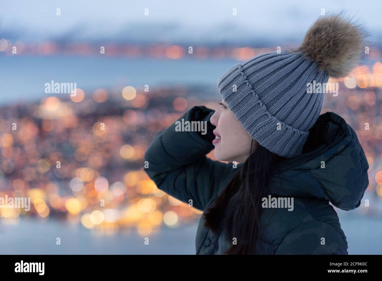 Aufgeregt junge Weibchen in khaki Daunenjacke und grau warm hut schaut weg und betrachten erstaunliche Winter Blick auf die Stadt Am Abend an der Küste gelegen Stockfoto