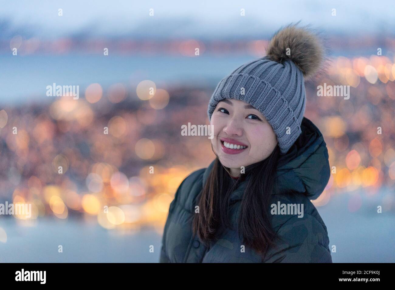 Aufgeregt junge Weibchen in khaki Daunenjacke und grau warm hat Blick auf die Kamera und erwägen erstaunliche Winter Blick auf Stadt an der Küste am Abend gelegen Stockfoto