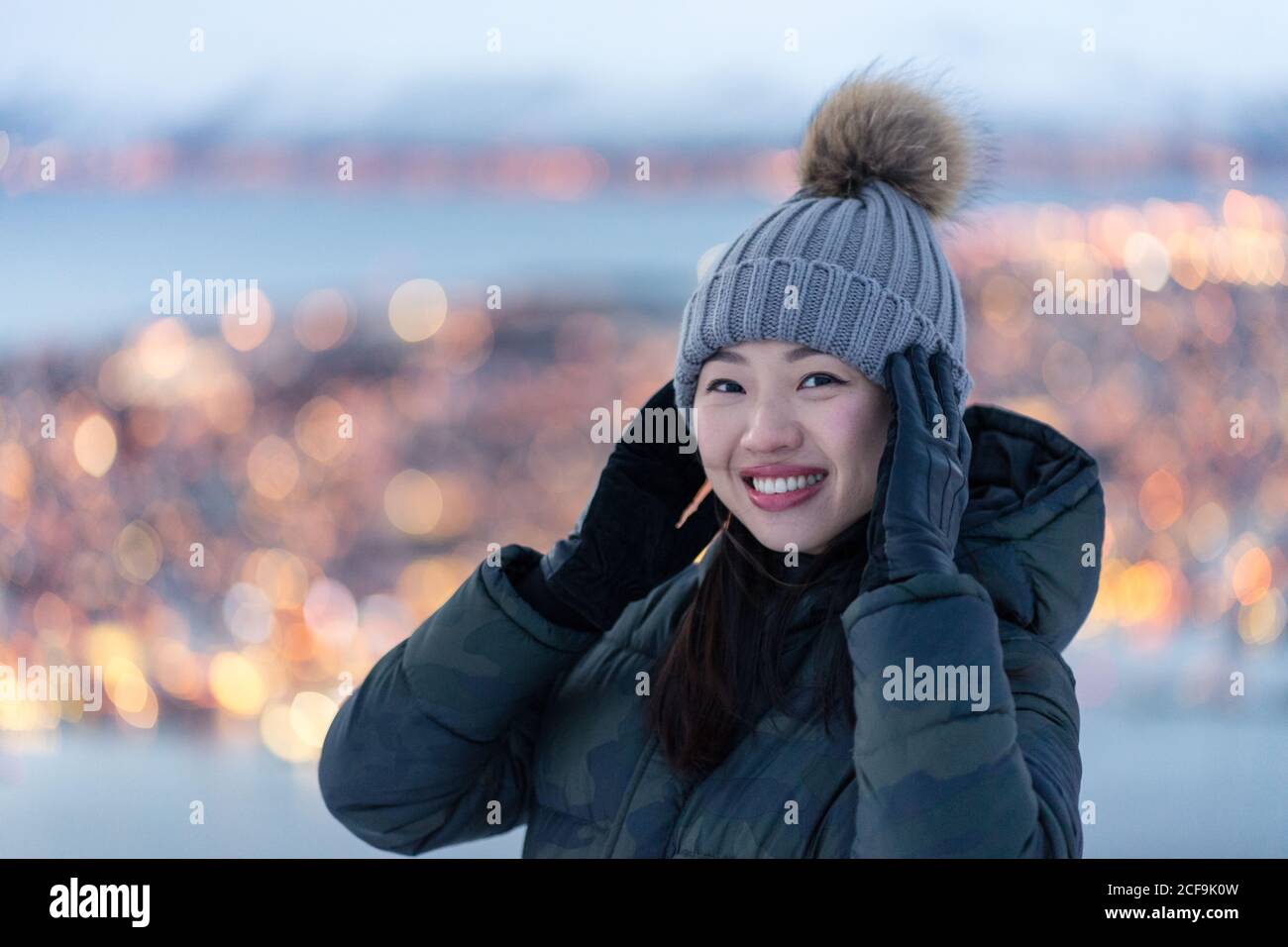 Aufgeregt junge Weibchen in khaki Daunenjacke und grau warm hat Blick auf die Kamera und erwägen erstaunliche Winter Blick auf Stadt an der Küste am Abend gelegen Stockfoto