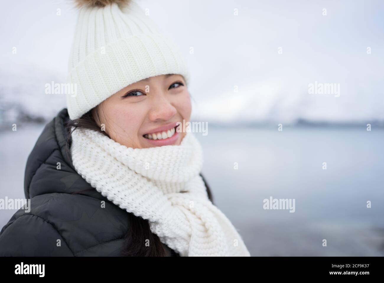 Glücklich Kamera weiblich mit erstaunlichen Augen in schwarzer Jacke und Weiße warme Mütze und Schal Blick auf Kamera und lächelnd Vor grauem verschwommenem Hintergrund in Norwegen Stockfoto