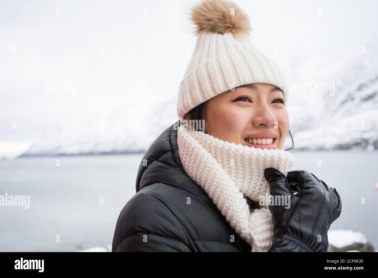 Glückliche Hündin mit erstaunlichen Augen in schwarzer Jacke und weiß Warmer Hut und Schal blicken weg und lächeln gegen Weiß Verschwommener Hintergrund in Norwegen Stockfoto