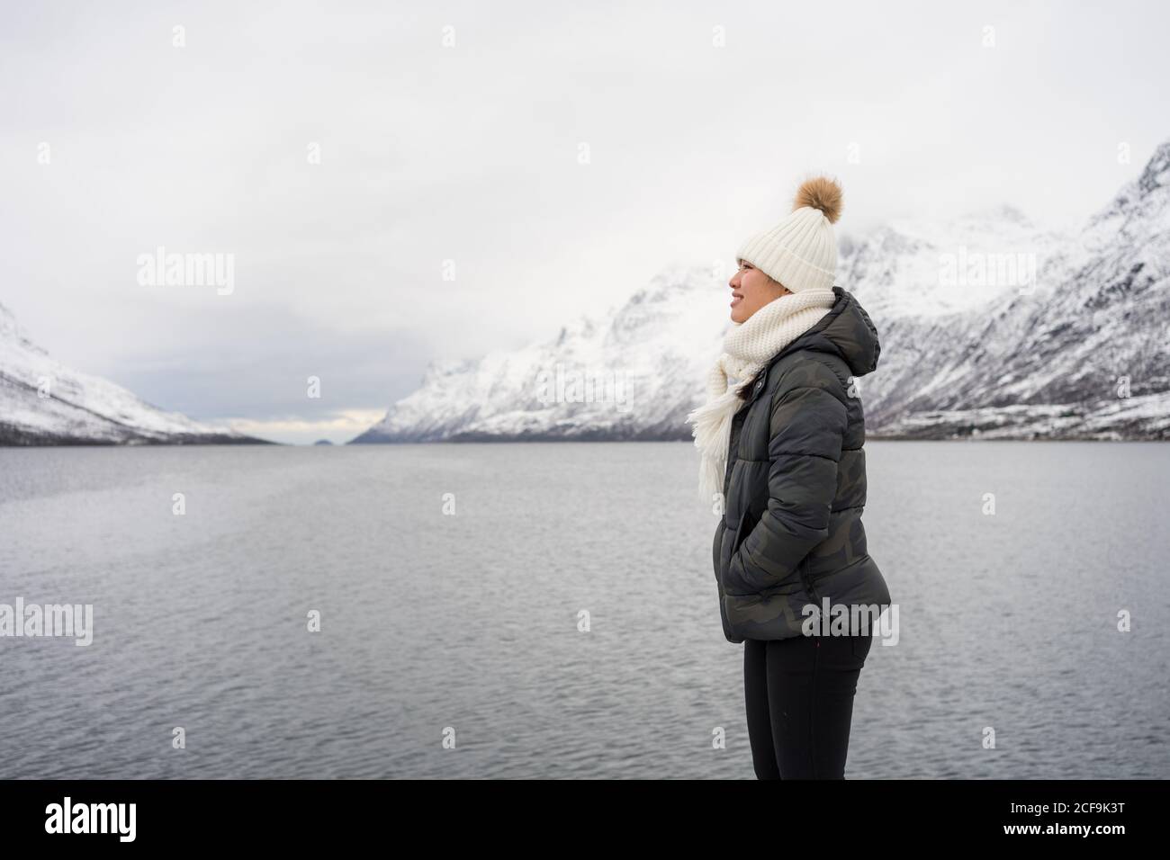 Glückliche Hündin mit erstaunlichen Augen in schwarzer Jacke und weiß Warmer Hut und Schal blicken weg und lächeln gegen Weiß Verschwommener Hintergrund in Norwegen Stockfoto