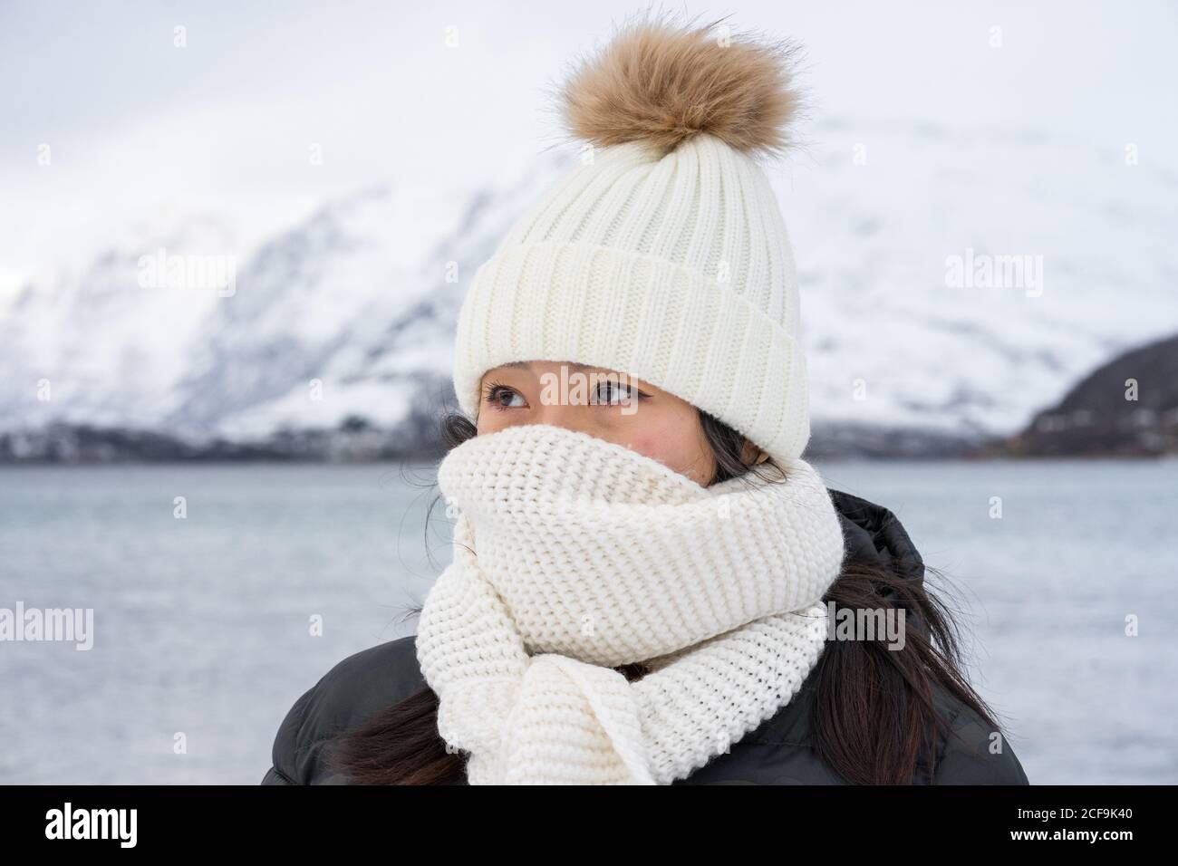 Junge Frau mit schwarzer Jacke und weißem warmen Hut und Schal, die vor grauem, unscharfem Hintergrund in Norwegen auf die Kamera schaut Stockfoto