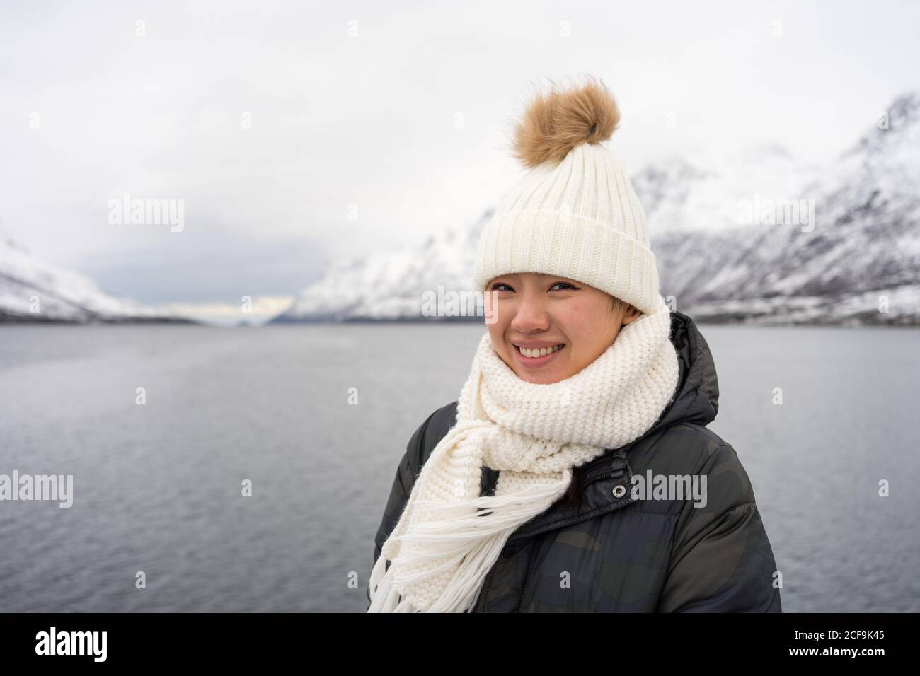 Glückliche Hündin mit erstaunlichen Augen in schwarzer Jacke und weiß Warmer Hut und Schal schauen auf die Kamera und lächeln dagegen Grau verschwommener Hintergrund in Norwegen Stockfoto