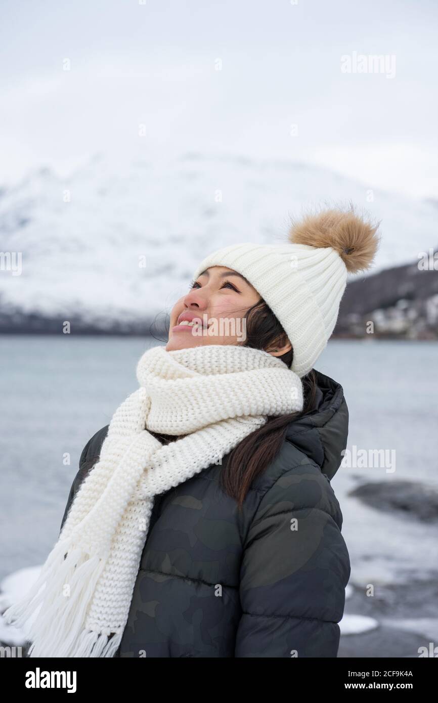 Glückliche Hündin mit erstaunlichen Augen in schwarzer Jacke und weiß Warmer Hut und Schal schauen nach oben und lächeln gegen grau Verschwommener Hintergrund in Norwegen Stockfoto