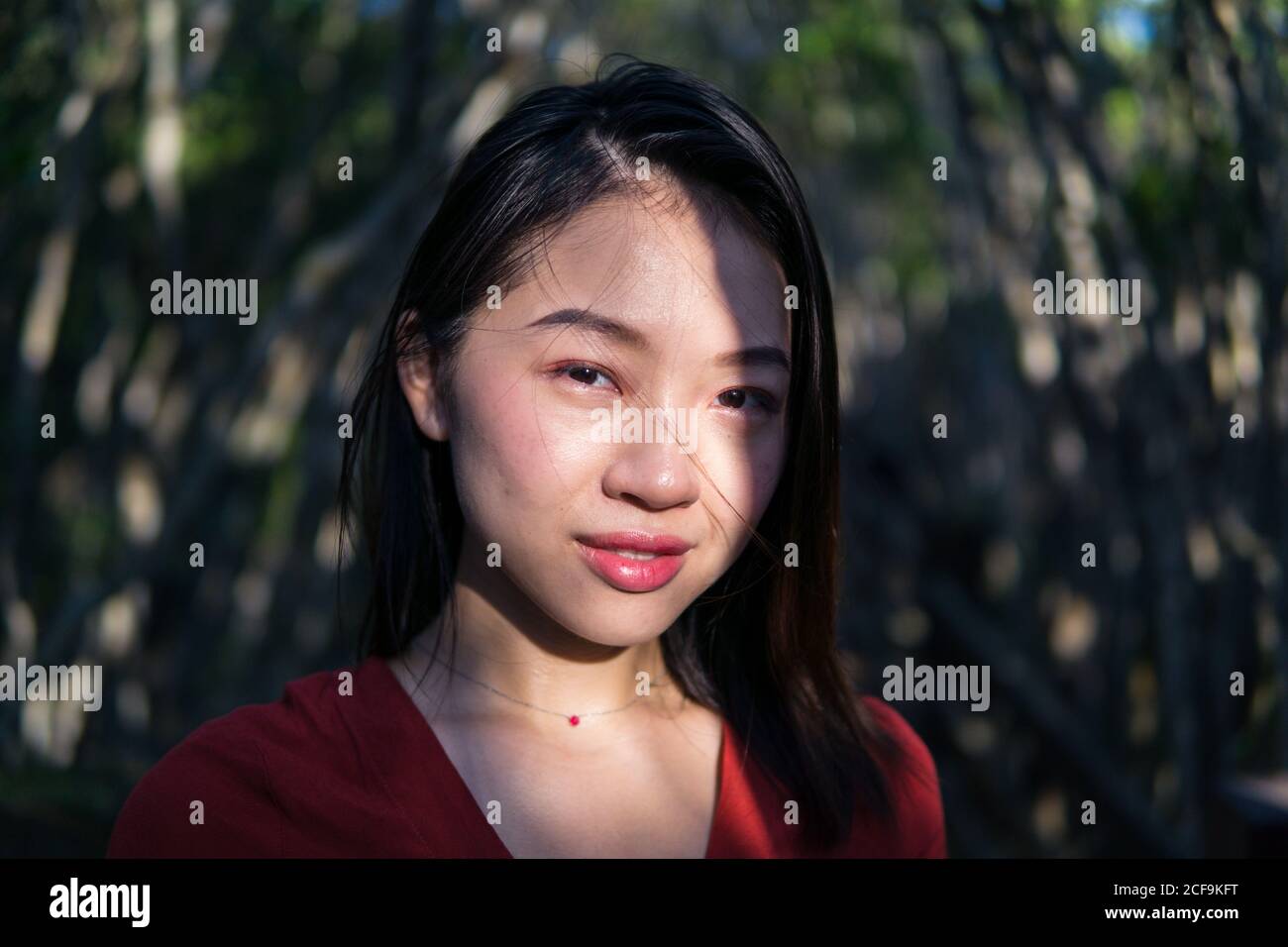 Nachdenklich junge asiatische Frau in Freizeitkleidung und kleine Halskette Blick auf die Kamera, während im Wald bei Tageslicht stehen Stockfoto