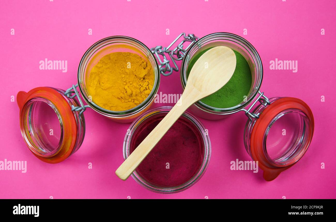 Draufsicht auf isolierte Einmachgläser und Holzlöffel mit gelbem Kurkuma, roter Rote Bete und grünem Spinatpulver, rosa Hintergrund. Stockfoto