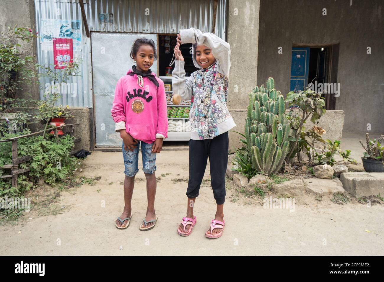 Maubisse, Osttimor - 10. AUGUST 2018: Glückliches ethnisches Mädchen, das Friedenszeichen zeigt, während es mit einem fröhlichen Freund neben einem lokalen Geschäft steht Stockfoto