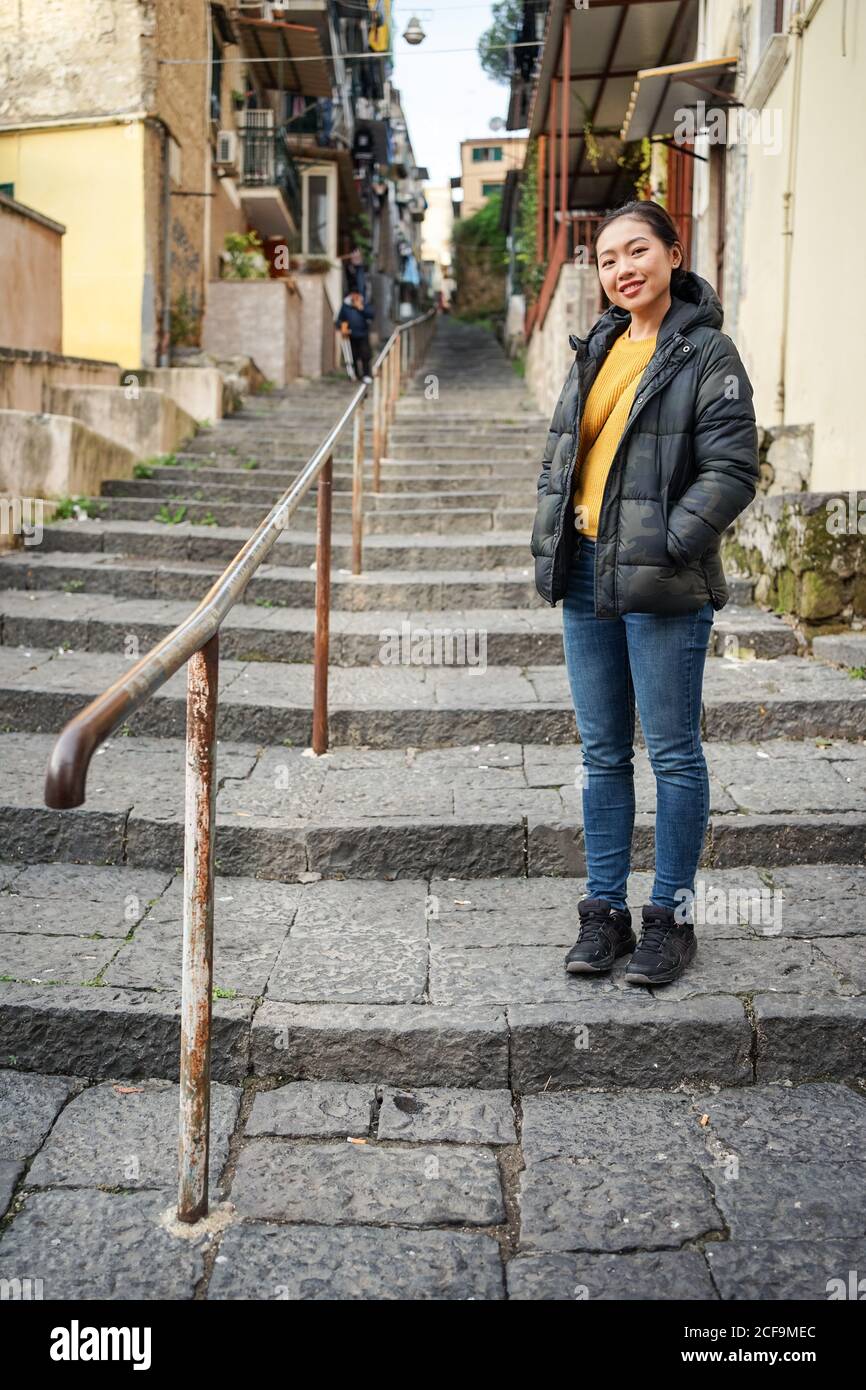 Ganzkörper asiatische weiblich mit Händen in Taschen Blick auf Kamera, während Sie auf Treppen vor verwitterten Gebäuden in der Stadt stehen Straße Stockfoto
