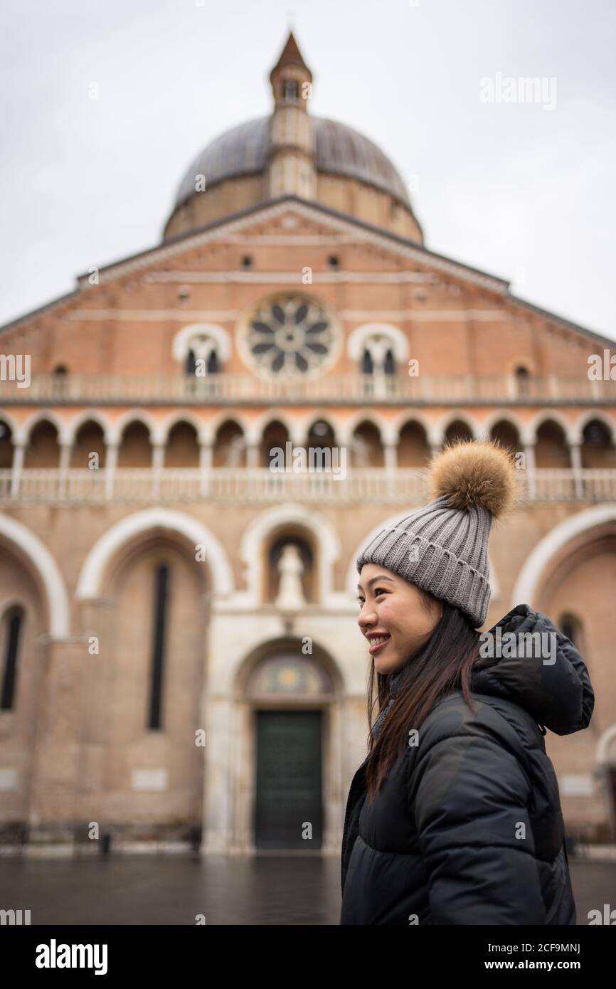 Seitenansicht einer angenehmen asiatischen Reisenden in warmer Kleidung Und Hut lächelnd und wegschauen mit alten großen Basilika Von San Antonio auf verschwommenem Hintergrund in Padova bei Italien Stockfoto