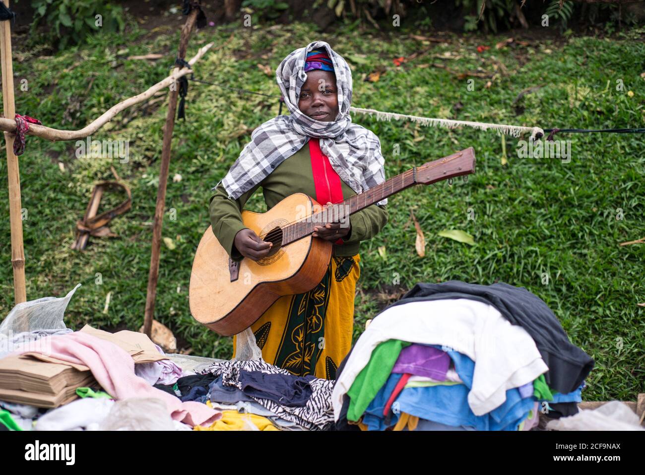 Ruanda, Afrika - 14. Dezember 2019: Ruhige afroamerikanische Frau in farbenfrohen einfachen Kleidern, die Gitarre spielen und Kleidung im Marktstand verkaufen und dabei die Kamera betrachten Stockfoto