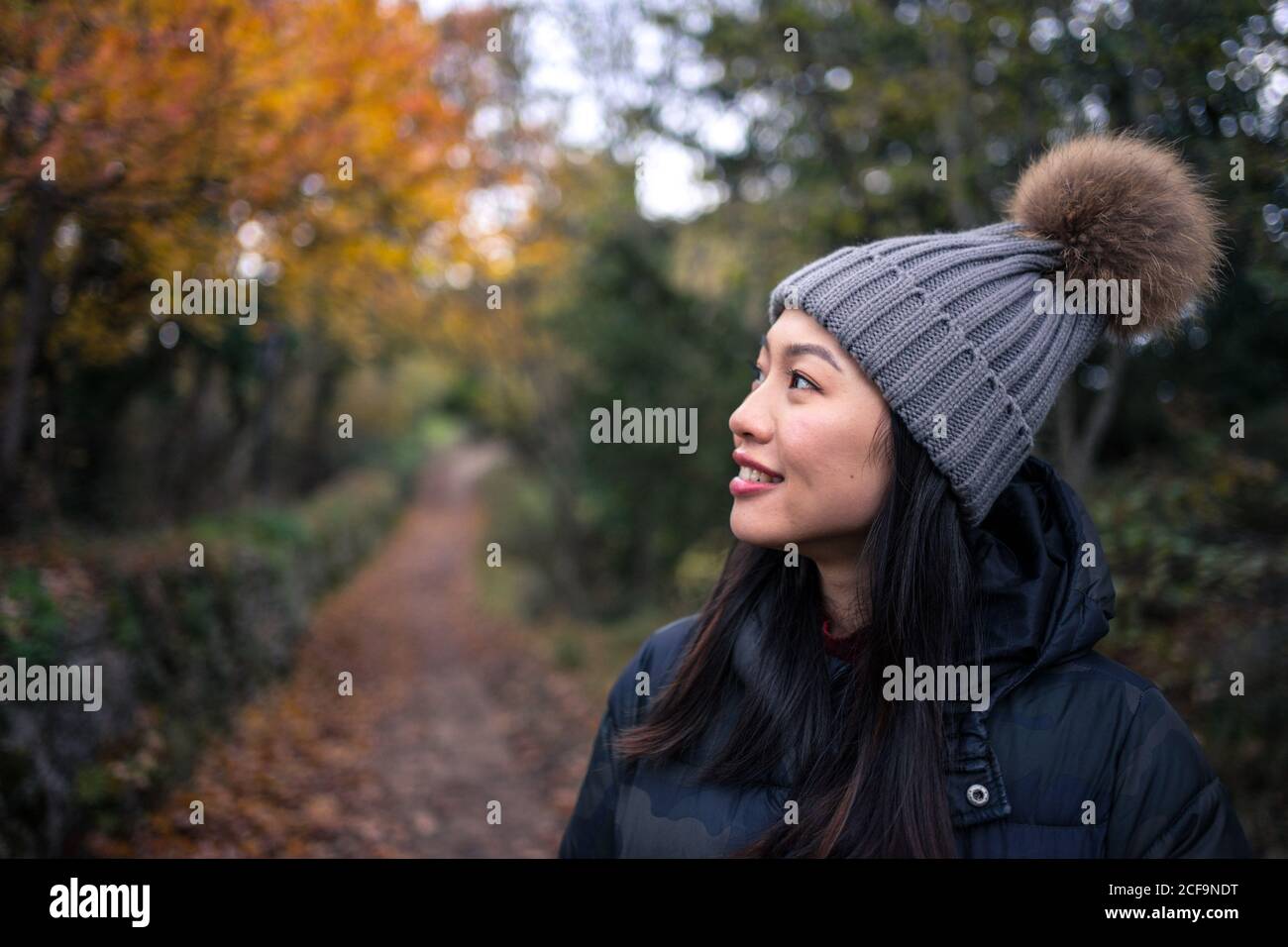 Junge charmante asiatische Frau in schwarzer warmer Jacke und Strickmütze mit Bommel lächelnd und mit Herbstgängern in San Marino, Italien, auf der Straße in den Garten blickend Stockfoto