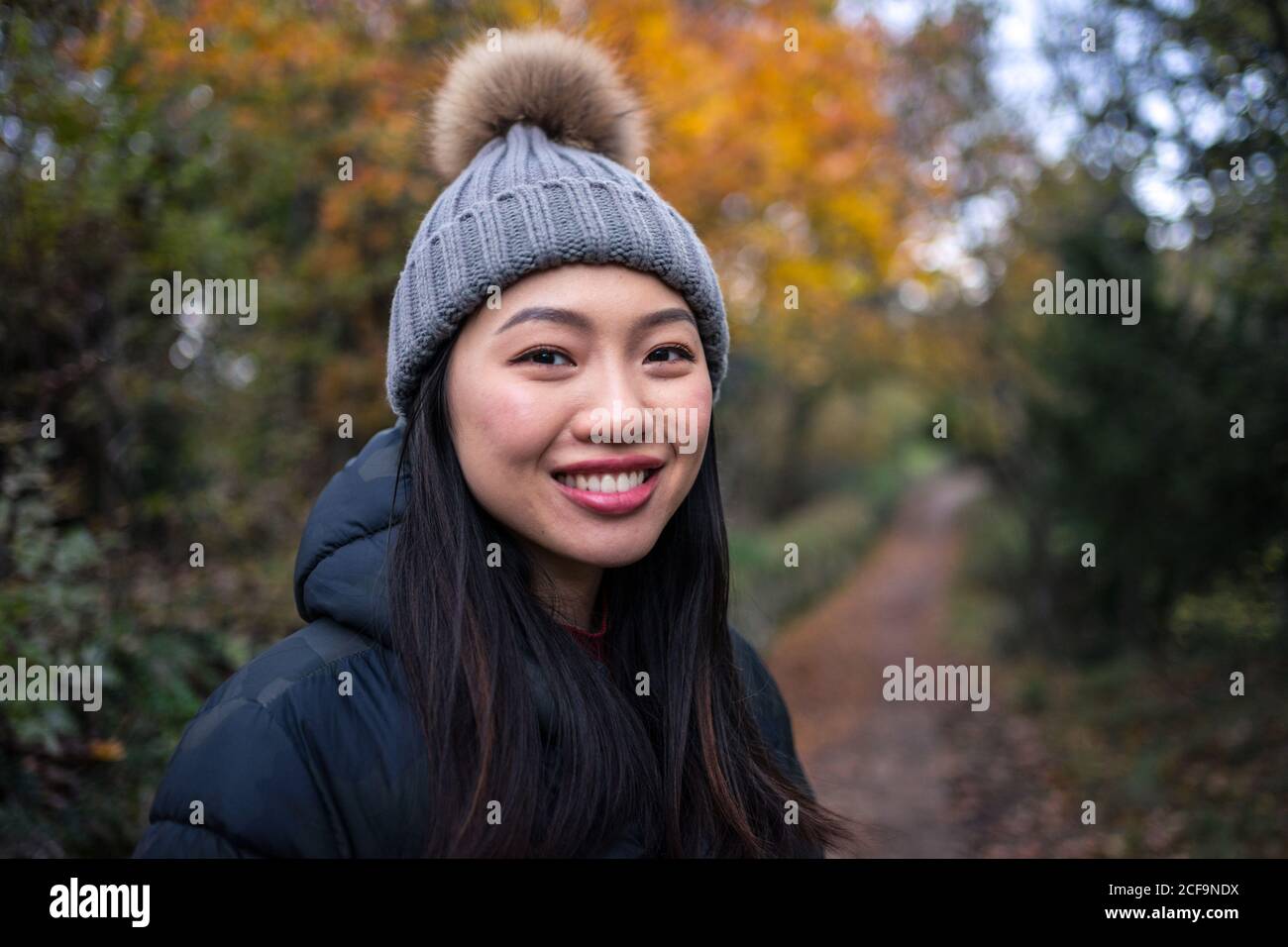 Junge charmante asiatische Frau in schwarzer warmer Jacke und Strickmütze mit Bommel lächelnd und Blick auf die Kamera auf dem Weg in den Garten mit Herbstgängern in San Marino, Italien Stockfoto