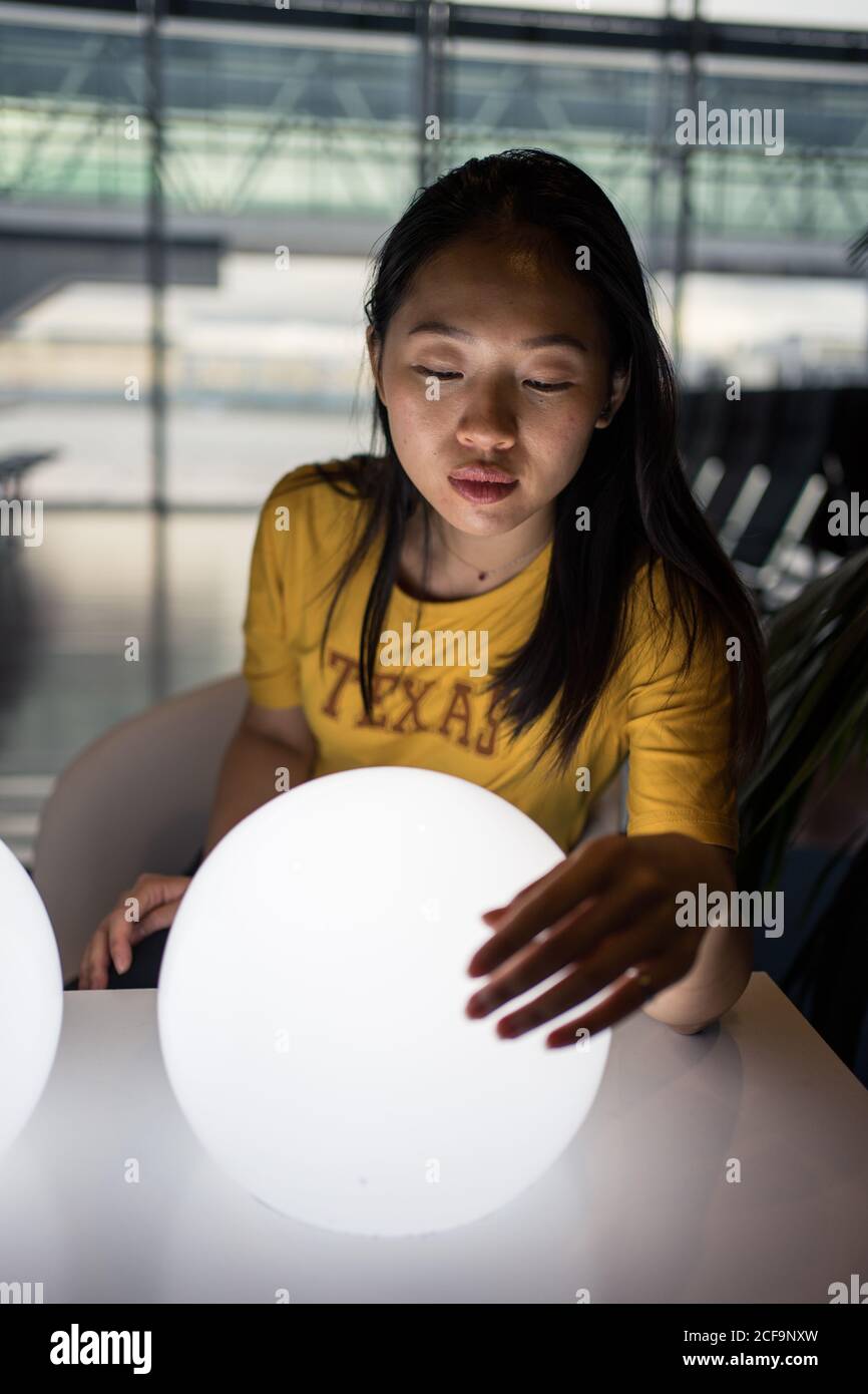 Langhaarige, ausgestrahlte asiatische Frau, die in der Wartehalle weiße, runde Lampen auf dem Tisch erhellte und berührte Stockfoto