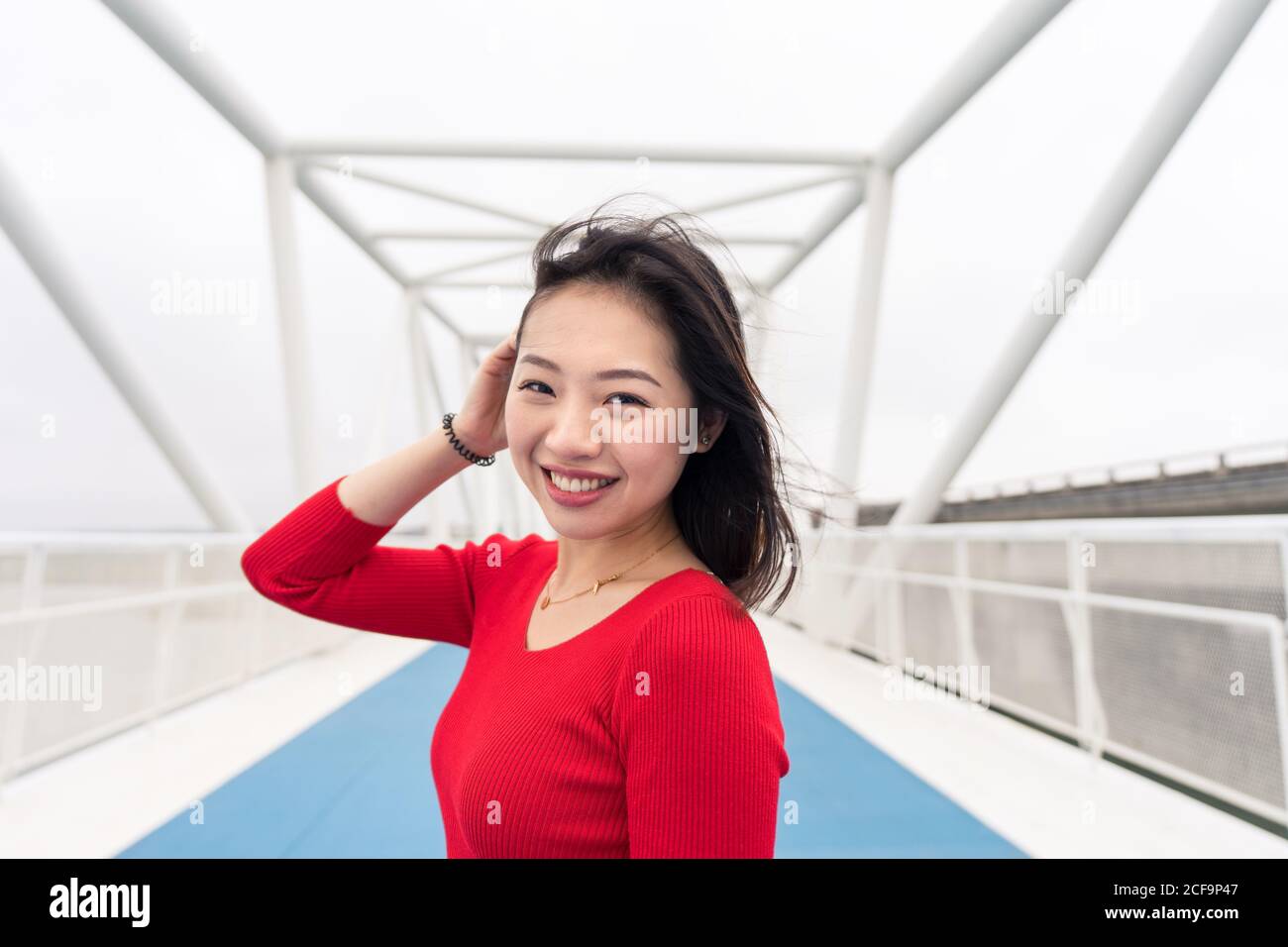 Charmante asiatische Dame im roten Hemd lächelnd und Blick auf Kamera beim Spaziergang auf der modernen Stadtbrücke Stockfoto