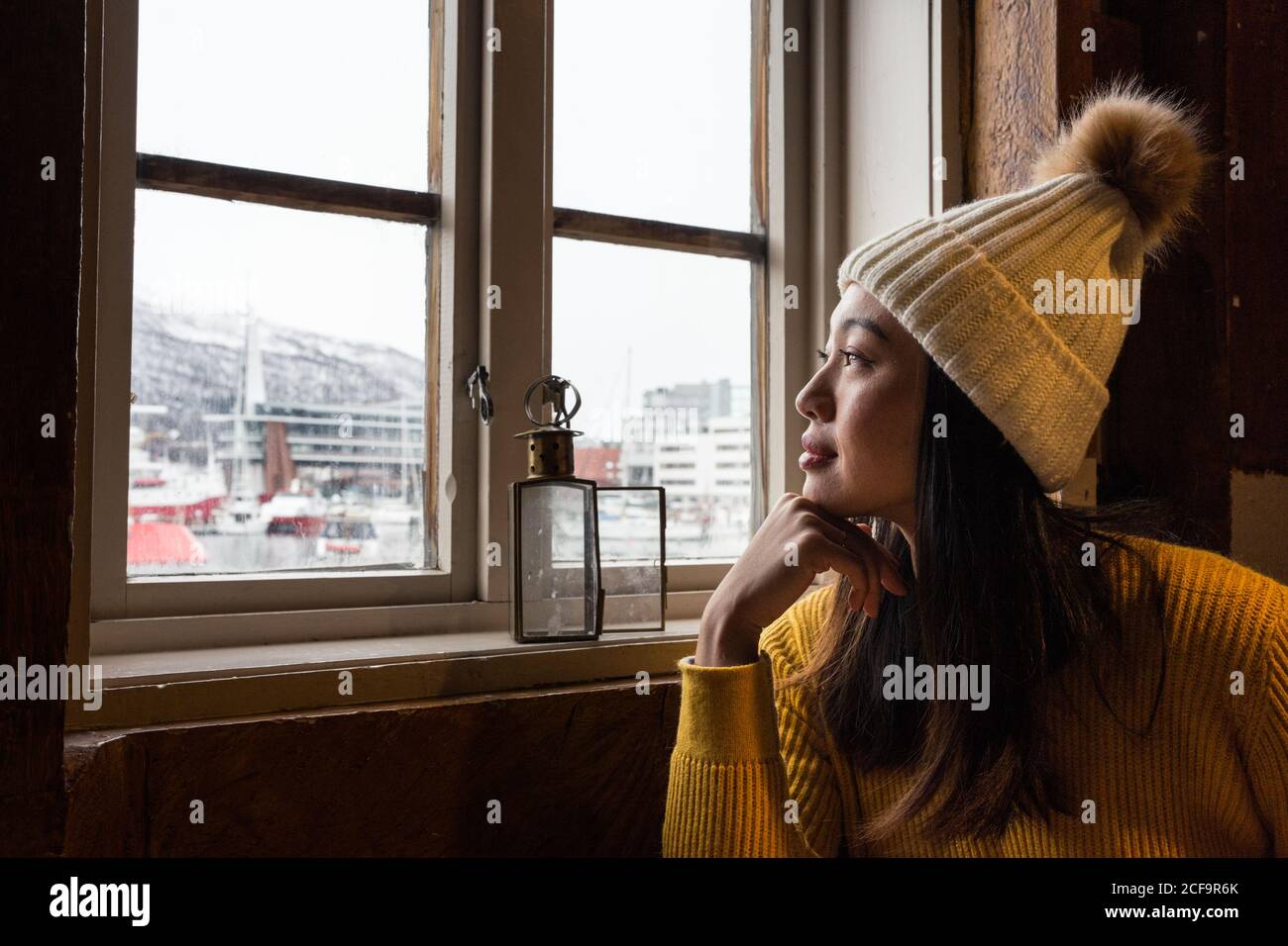 Seitenansicht der asiatischen nachdenklichen Frau im Urlaub in warm hut und gelber Pullover aus dem Fenster Tromso in Norwegen Stockfoto