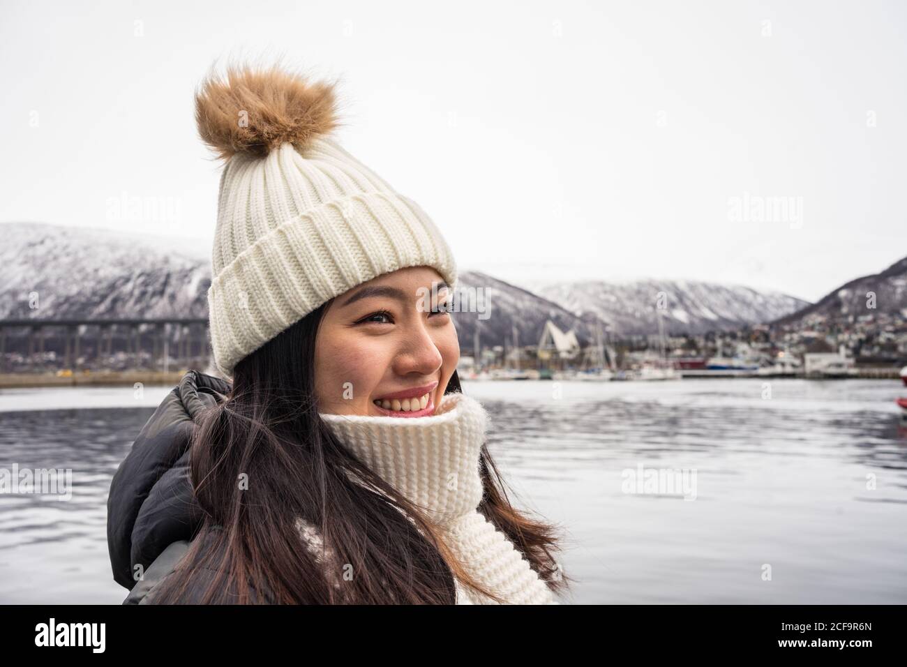 Junge fröhliche asiatische weibliche Tourist in warmer Kleidung und Hut Lächelnd beim Sightseeing mit Bergen und Meer im Hintergrund Tromso in Norwegen Stockfoto
