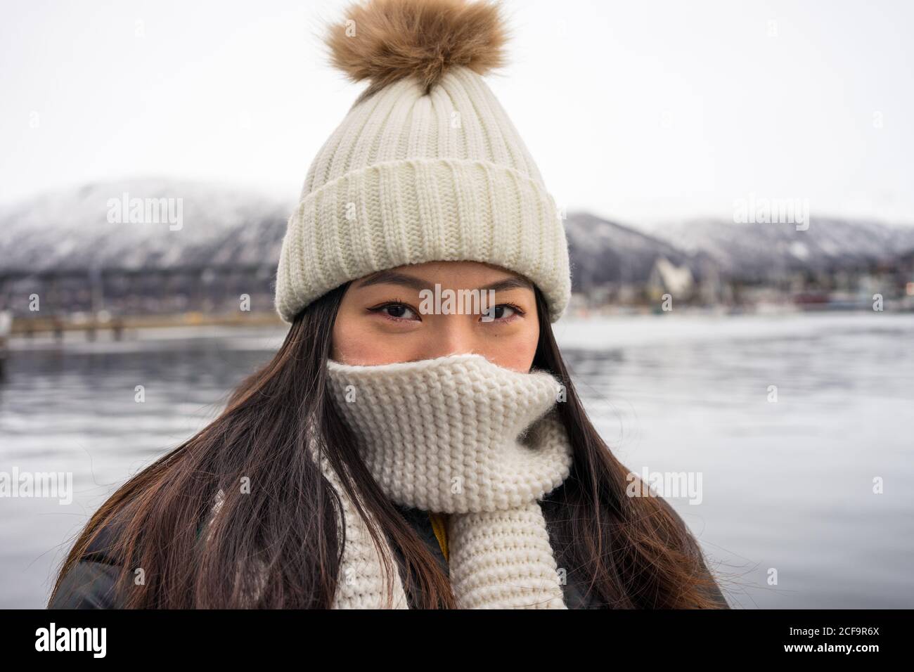 Junge fröhliche asiatische weibliche Tourist in warmer Kleidung und Hut Lächelnd beim Sightseeing mit Bergen und Meer im Hintergrund Tromso in NorwayBlick auf die Kamera Stockfoto