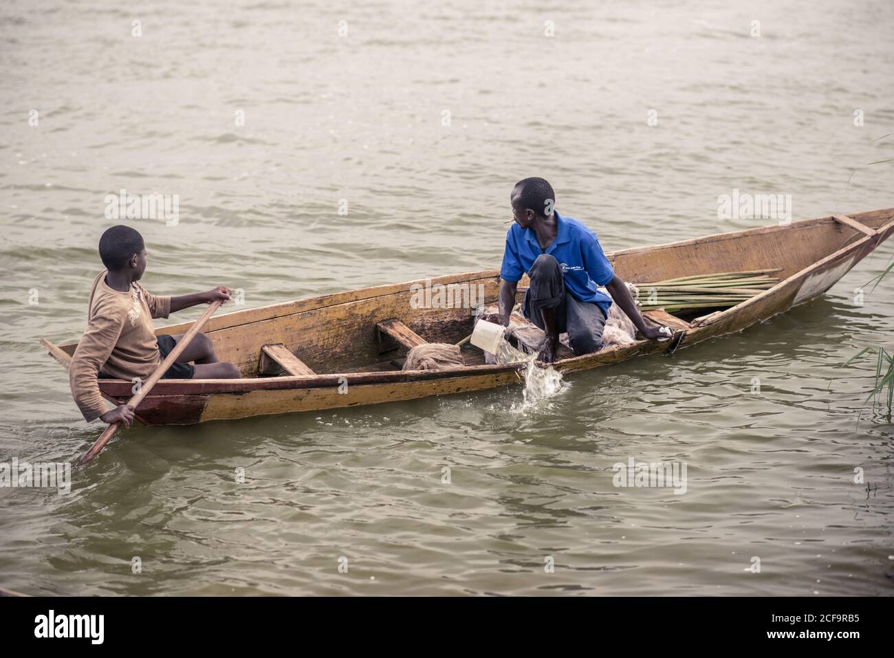 Uganda - November, 26 2016: Männlicher Teenager rudert, während erwachsener Mann Wasser aus dem alten Boot ausbackt und während der Fahrt auf dem schmutzigen Fluss wegschaut Stockfoto