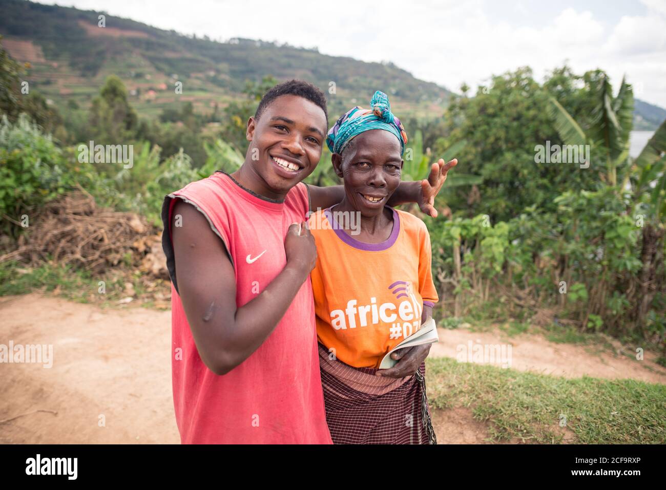 Uganda - November, 26 2016: Freundlicher schwarzer Mann umarmt ältere Frau und schaut auf die Kamera, während er auf Gras vor dem Dorf steht Stockfoto