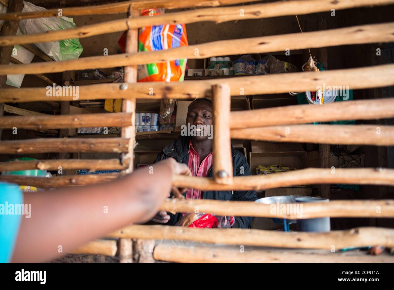 Uganda - 26. November 2016: Lächelnder nachdenklicher afrikanischer Verkäufer, der die Bestellung für einen nicht erkennbaren Kunden in lebendigen Kleidern fokussiert und vorbereitet, während er bei Tageslicht an der Theke mit frischem Gemüse und Waage im rustikalen Holzmarktstand steht Stockfoto