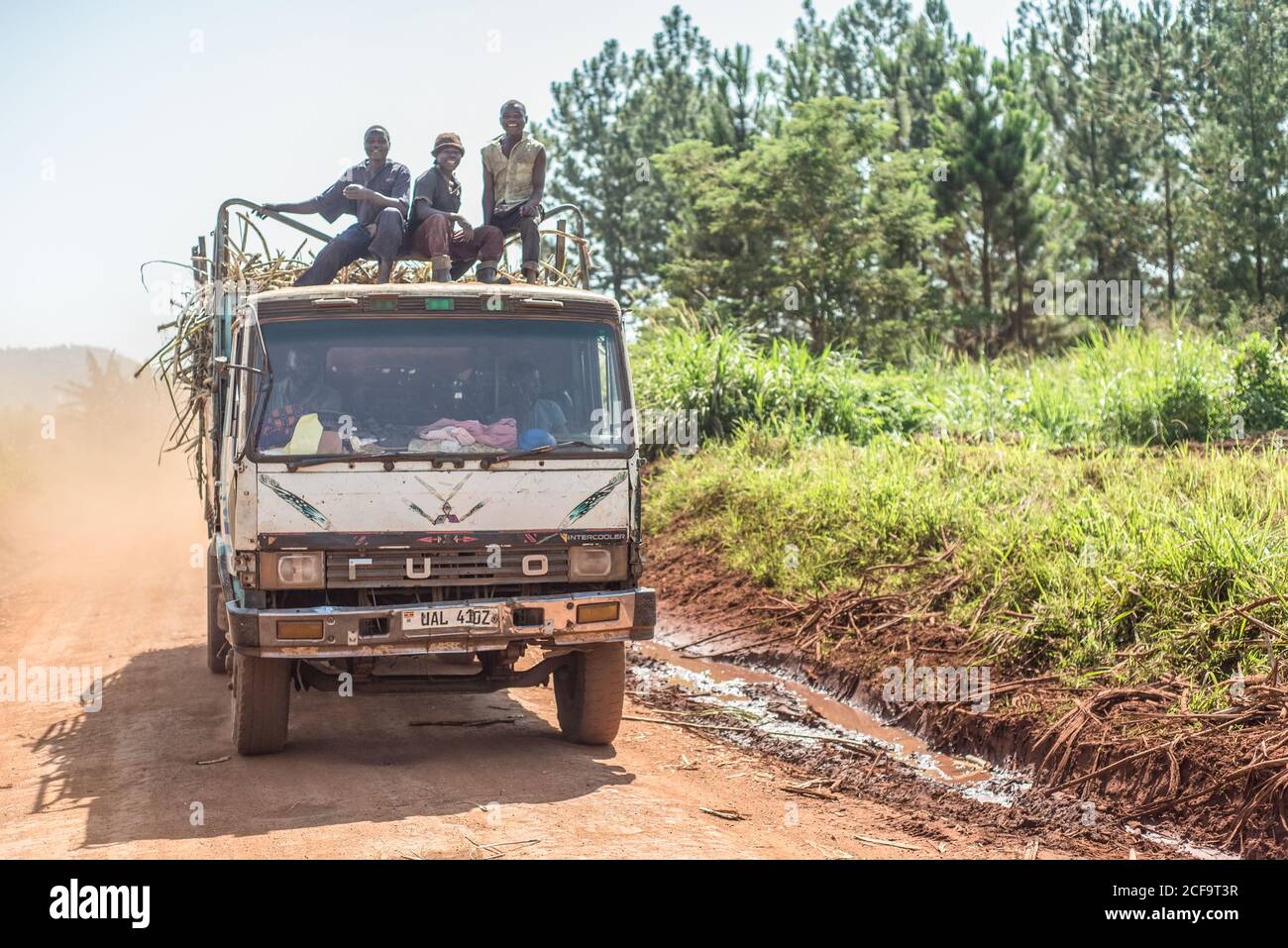 Uganda - 26. November 2016: Aufgeregte afrikanische Arbeiter sitzen auf dem Trailerzaun auf dem Kabinendach und lächeln vor der Kamera, während ein Mitarbeiter an heißen sonnigen Tagen mit einem LKW Fracht auf unbefestigten Straßen transportiert Stockfoto