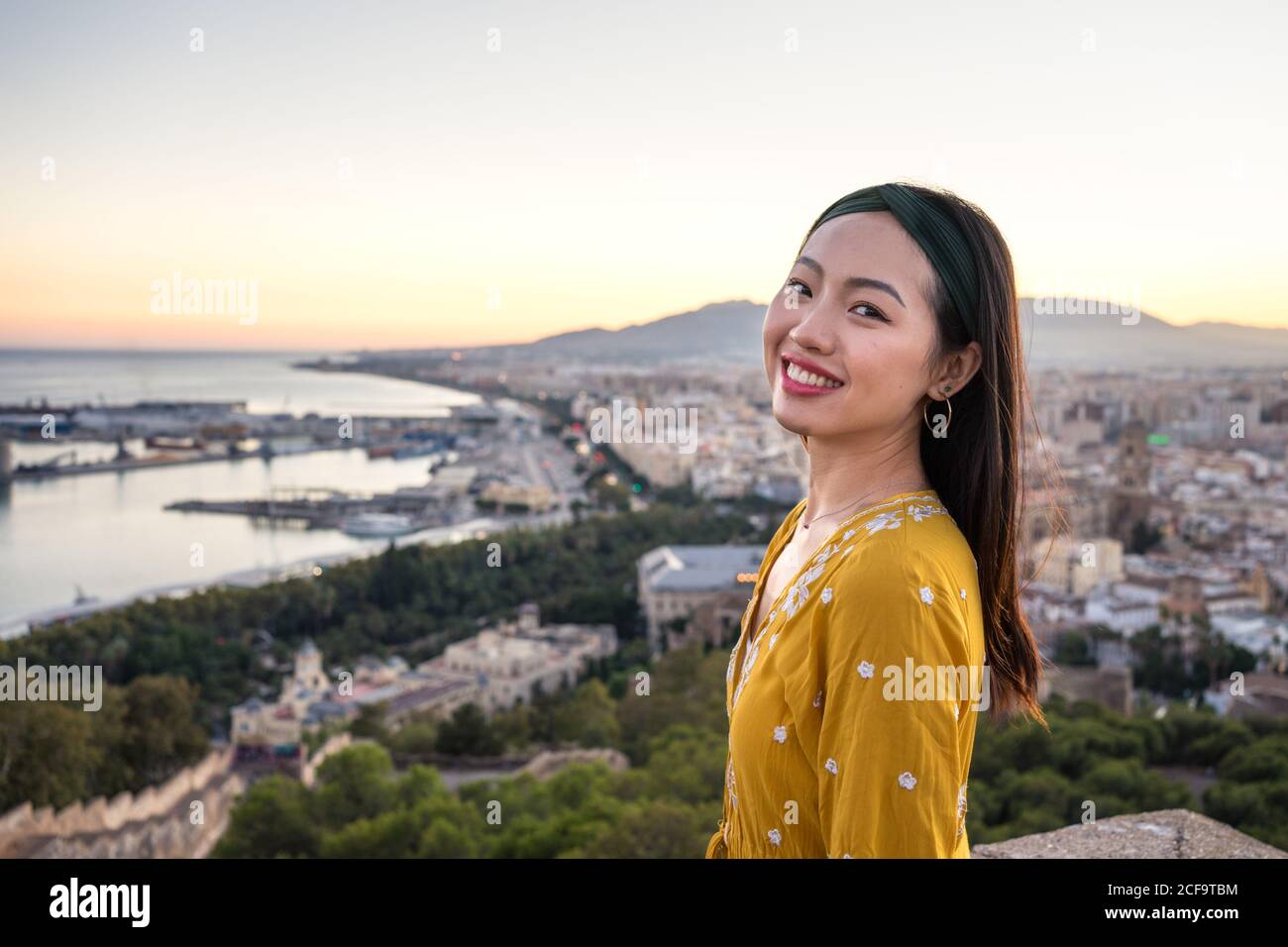 Seitenansicht einer optimistischen asiatischen Dame, die lächelt und die Kamera anschaut, während sie Castillo Gibralfaro auf dem verschwommenen Hintergrund der Küstenstadt und des Sonnenuntergartens in Malaga, Spanien besucht Stockfoto