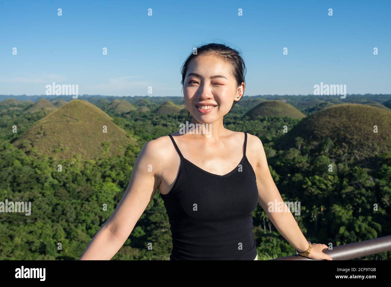 Fröhliche junge asiatische weibliche Tourist in schwarz oben und grau Hosen, die auf die Kamera blicken und dabei ein Auge blinzeln, während sie als Nächstes stehen Zum Zaun auf Aussichtsplattform mit grünen Hügeln und blau Himmel im Hintergrund Stockfoto
