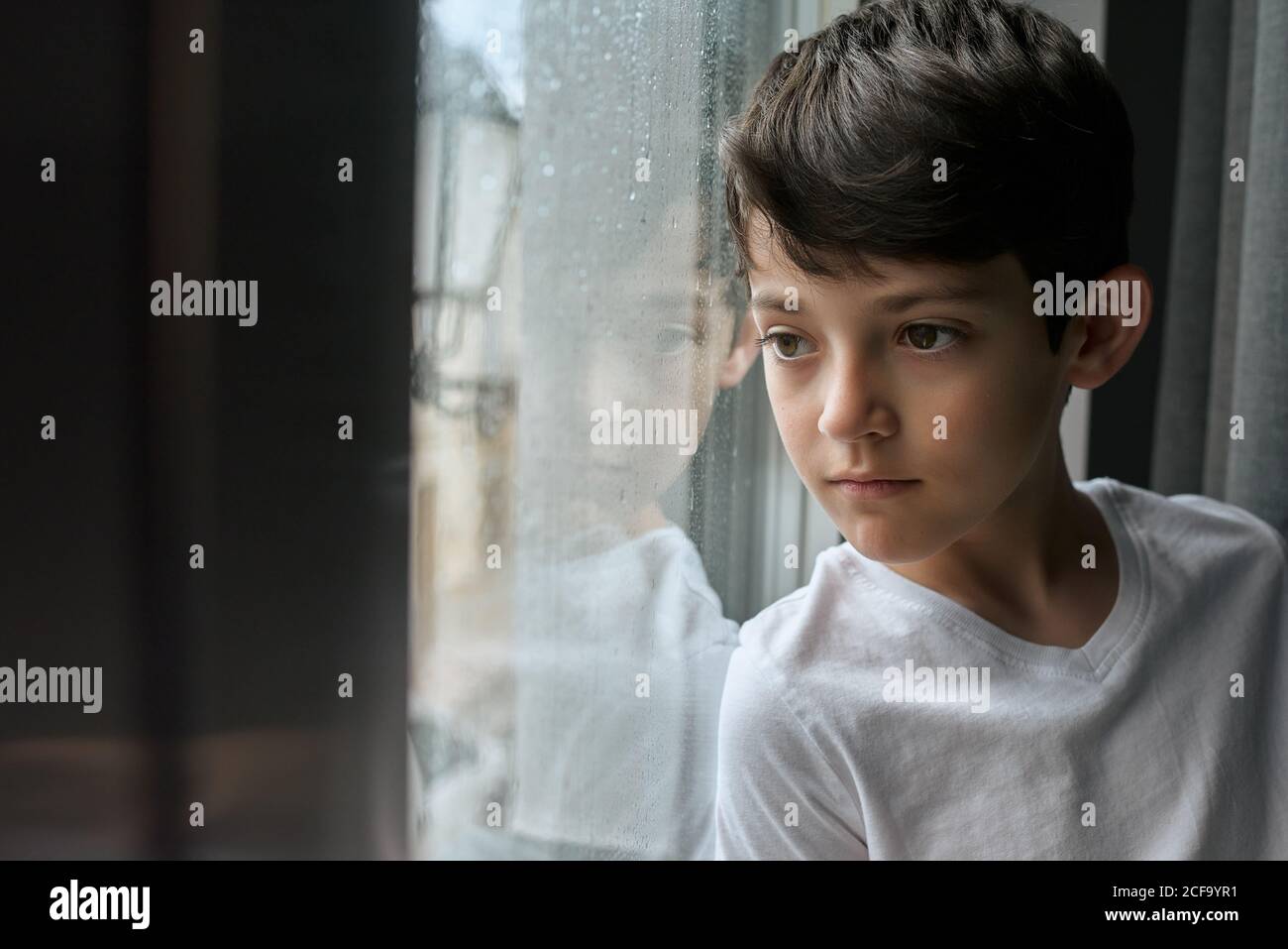 Gelangweiltes Kind in lässiger Kleidung im Wohnzimmer und stehen Lehnt sich an das Fenster und beobachtet den Regen auf der Straße Stockfoto