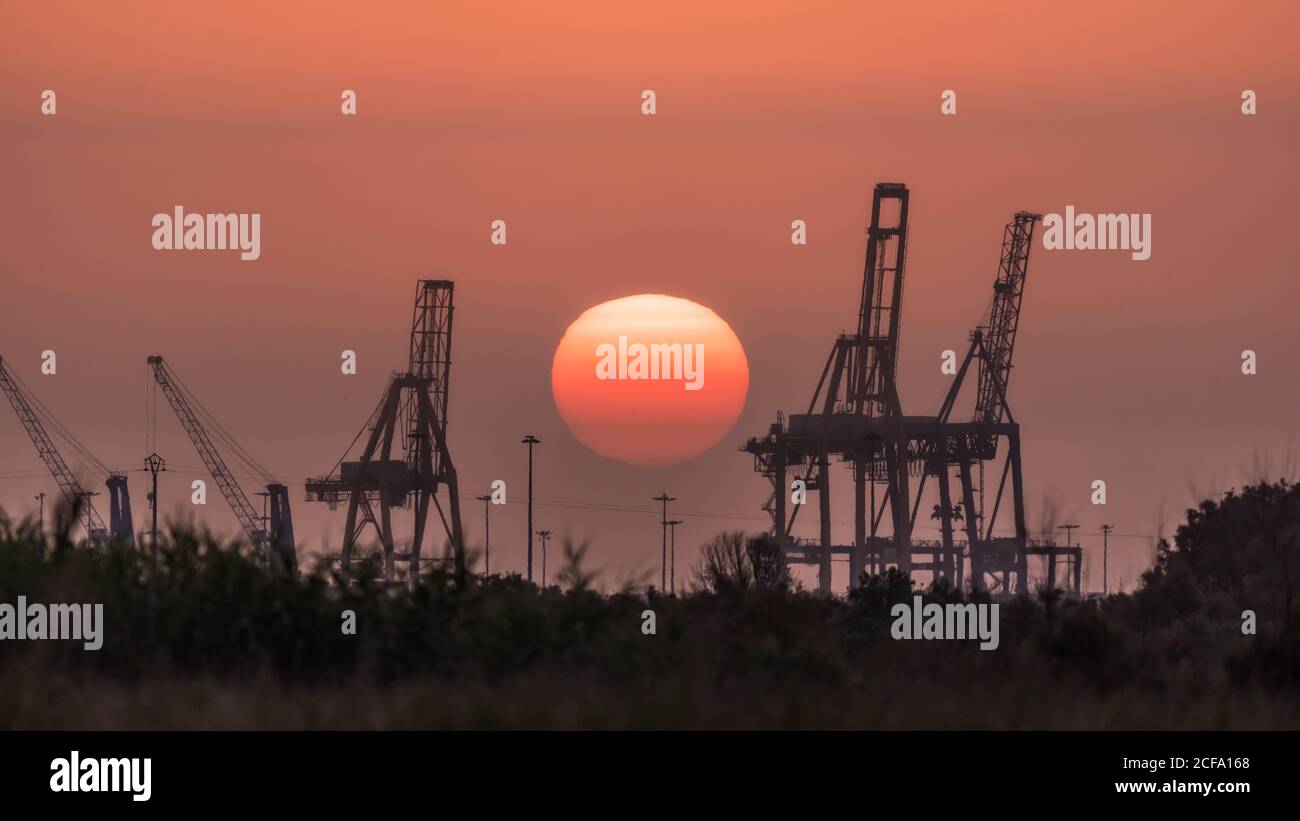 Silhouetten von modernen Baukräne gegen wolkenlosen Sonnenuntergang Himmel mit Große rote Sonne am Sommerabend Stockfoto