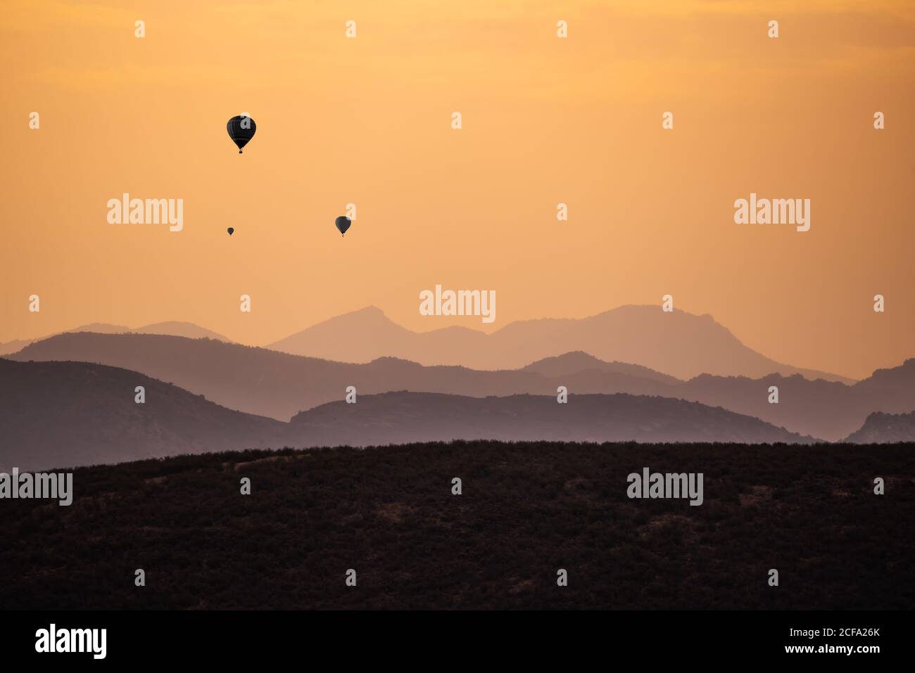 Silhouetten entfernter Heißluftballons, die gegen den Sonnenuntergang fliegen Über hügeliges Gelände Stockfoto